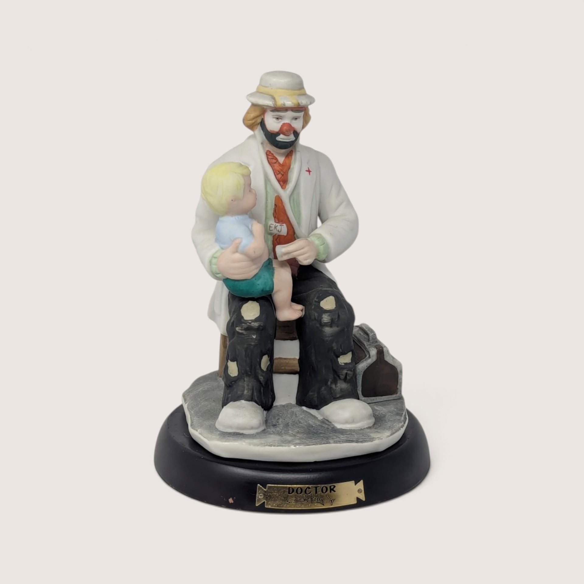 A vintage Flambro Emmett Kelly Jr. porcelain doctor figurine featuring a clown holding a child on a wooden base.
