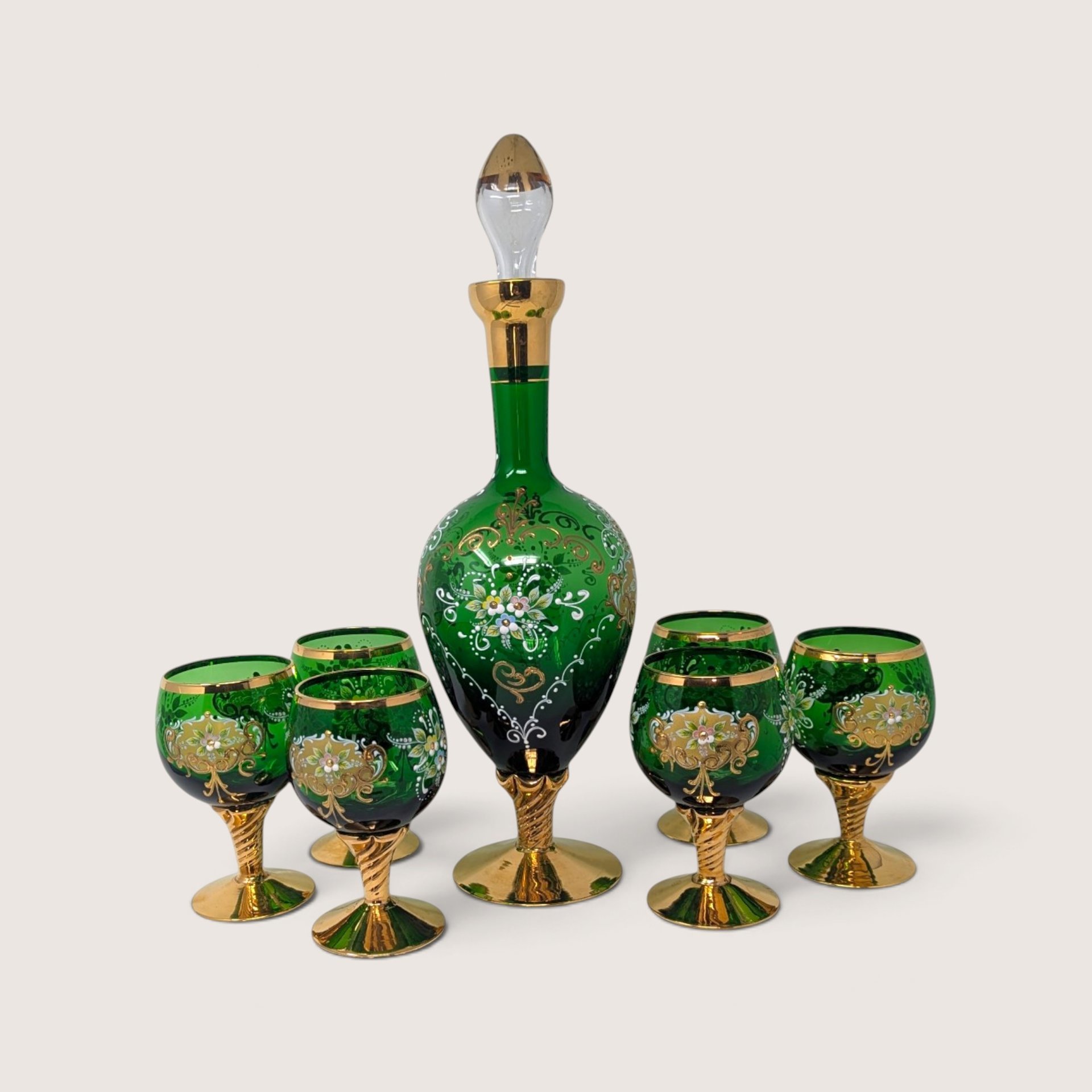 A vintage Italian emerald green glass decanter with stopper and six matching stemmed cordial glasses, all adorned with hand-painted white and gold floral enamel.