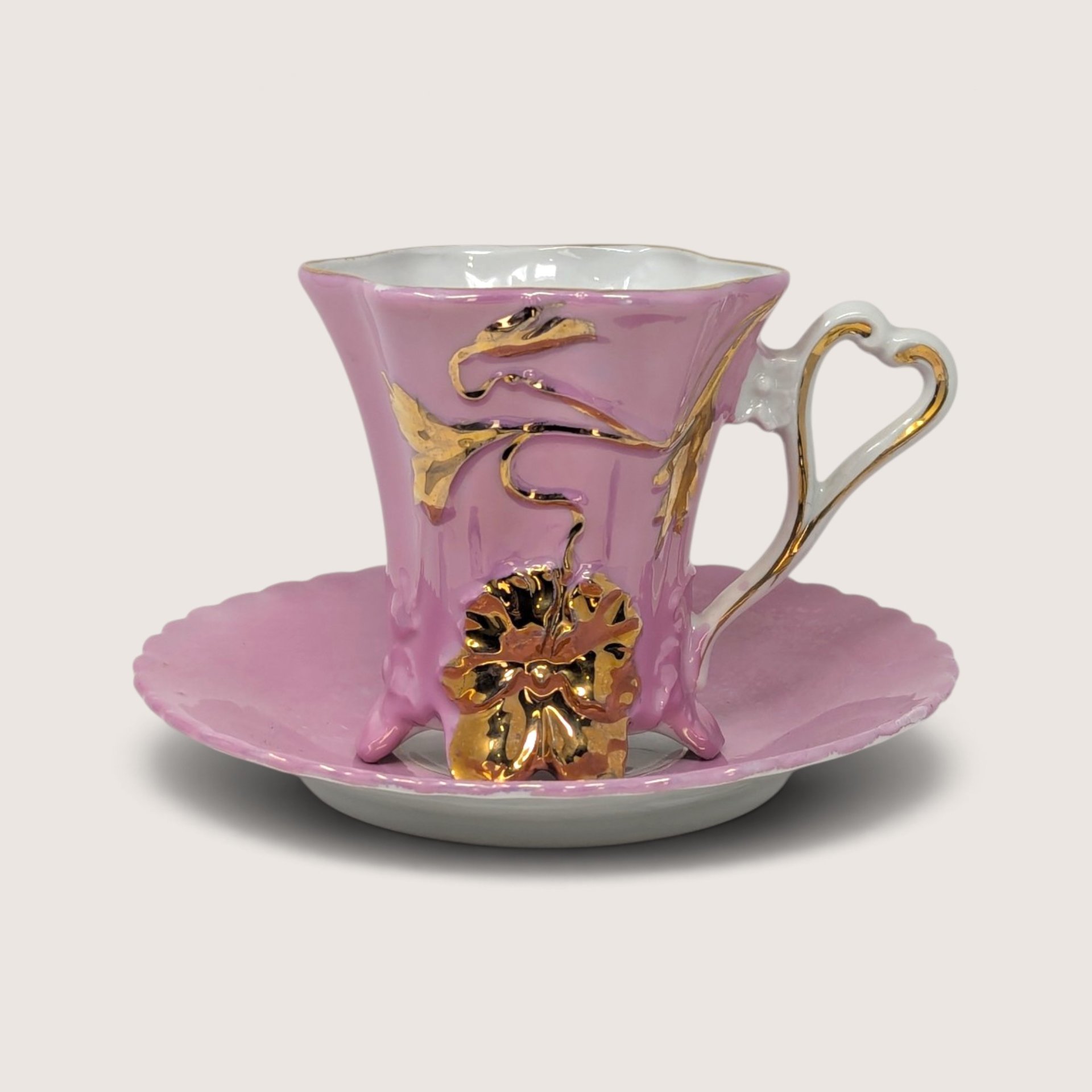 A vintage pink glazed ceramic footed teacup and matching saucer adorned with raised gold floral decorations and an ornate handle.