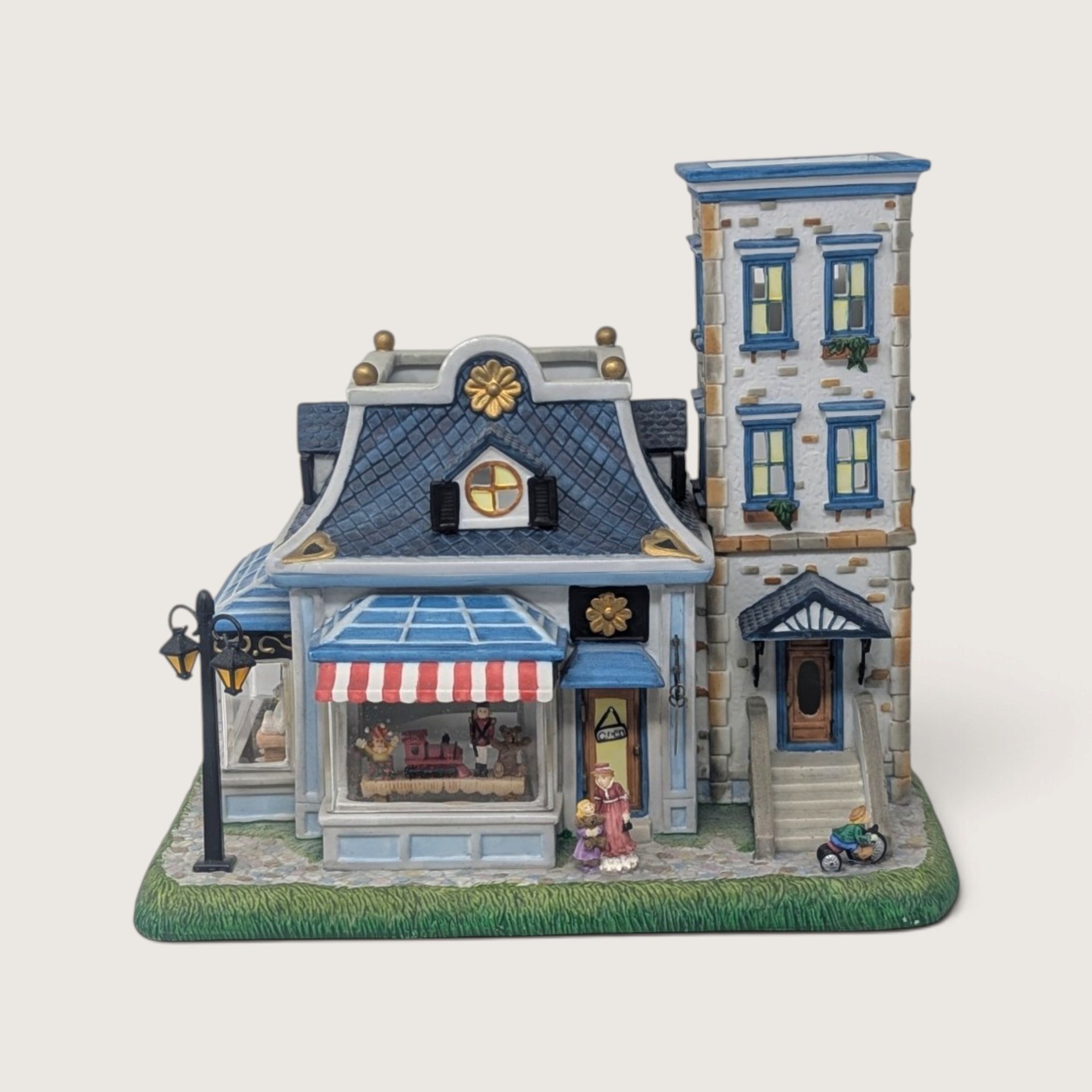 A vintage PartyLite Old World Village ceramic toy shoppe building with blue roofs, a striped awning, and cut-out windows against a neutral background.