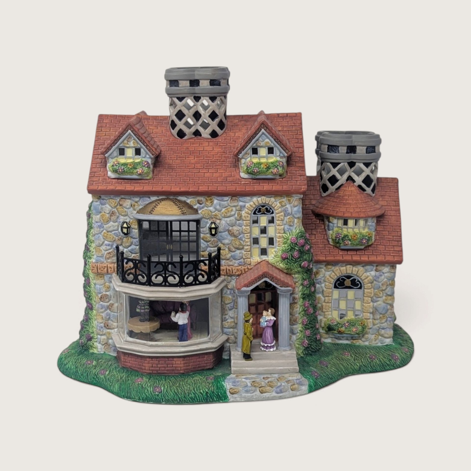 A vintage PartyLite Old World Village ceramic building featuring stone walls, a red shingle roof, a bay window, and figurines on the front steps.