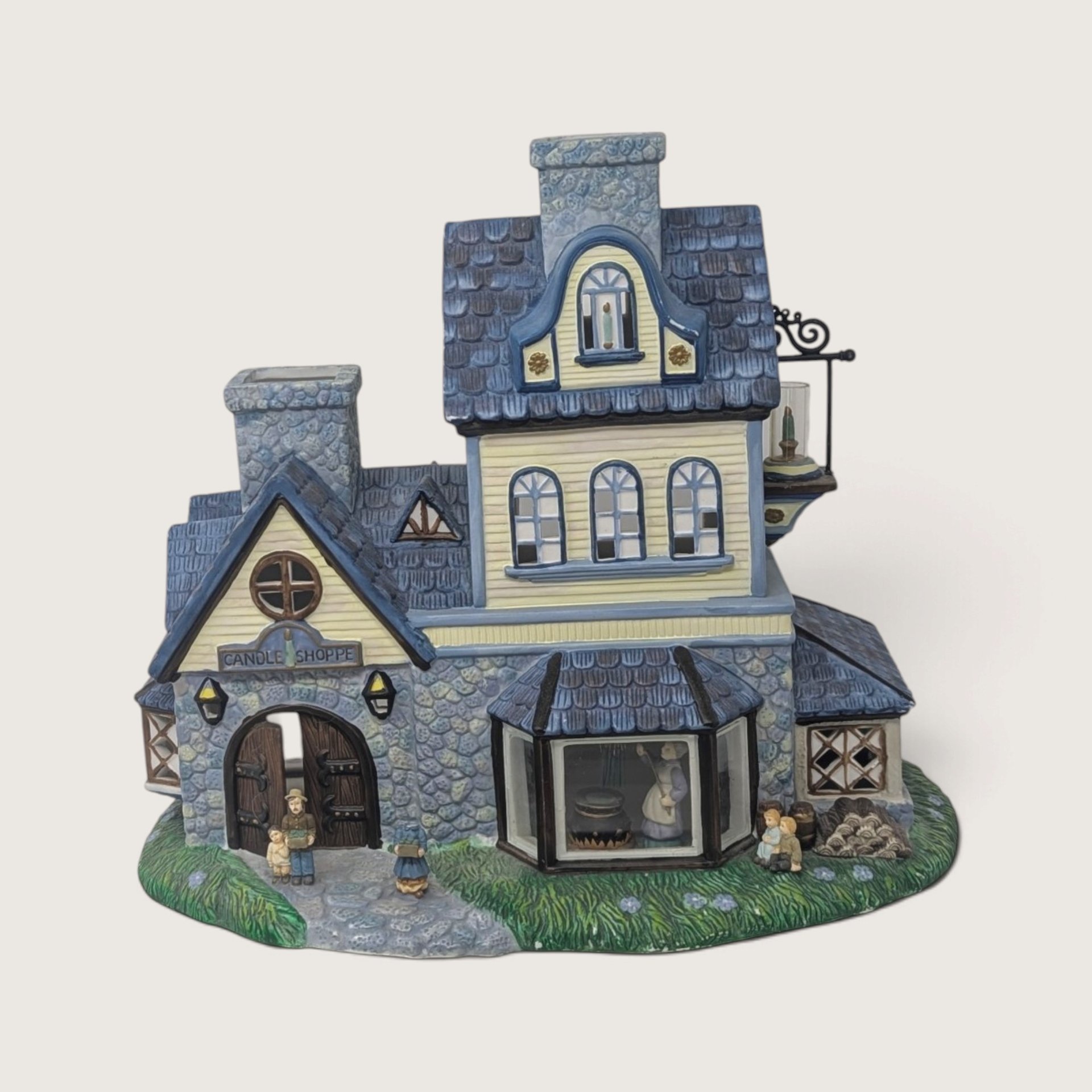 A vintage PartyLite Old World Village ceramic candle shoppe building featuring blue shingle roofs, yellow siding, stone accents, and a hanging lantern.