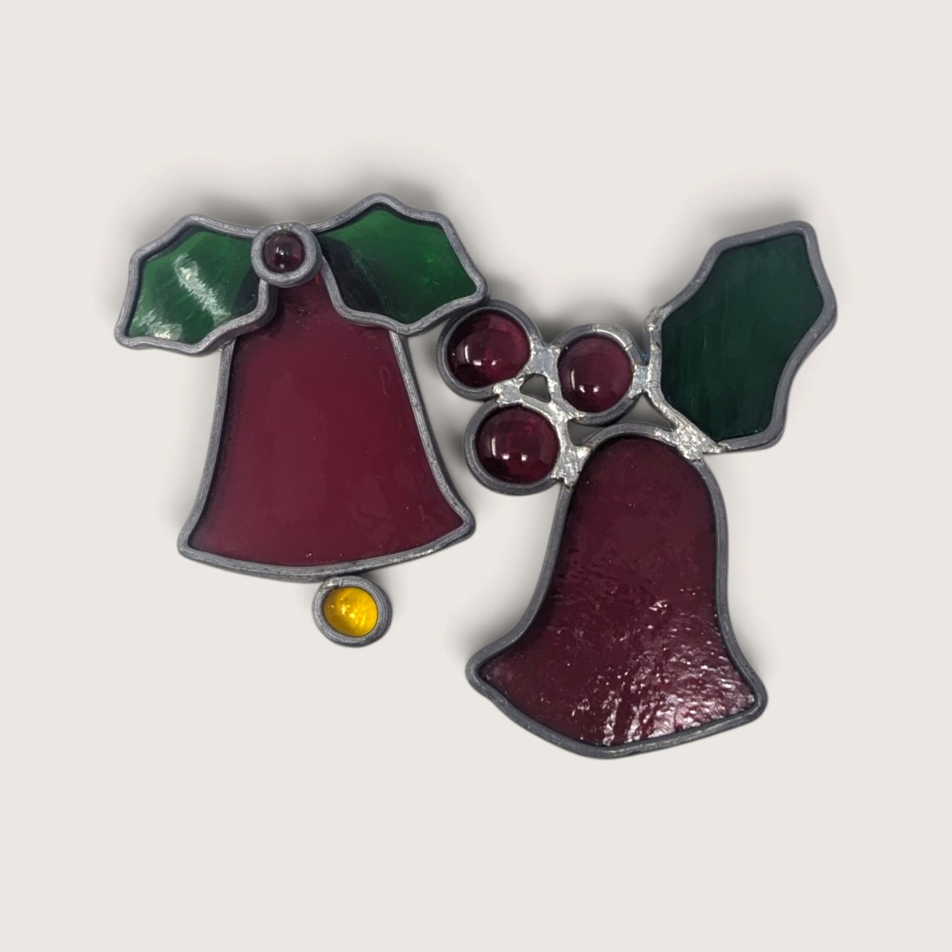 Two vintage stained glass Christmas ornaments shaped like bells with green leaves and red glass cabochons against a neutral background.