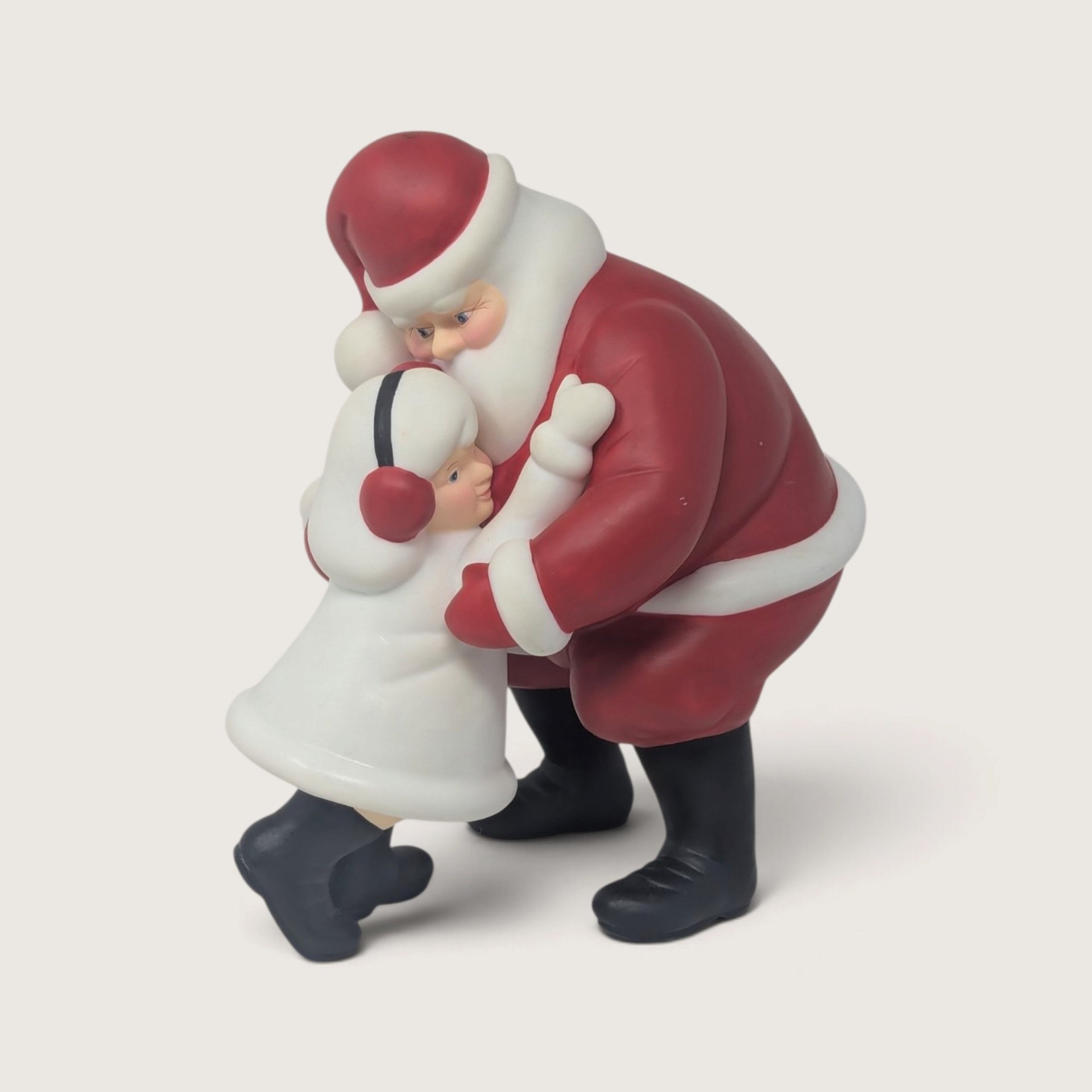 A vintage Department 56 Winter Silhouette ceramic figurine of Santa Claus in a red suit leaning down to embrace a child in a white coat with red earmuffs.