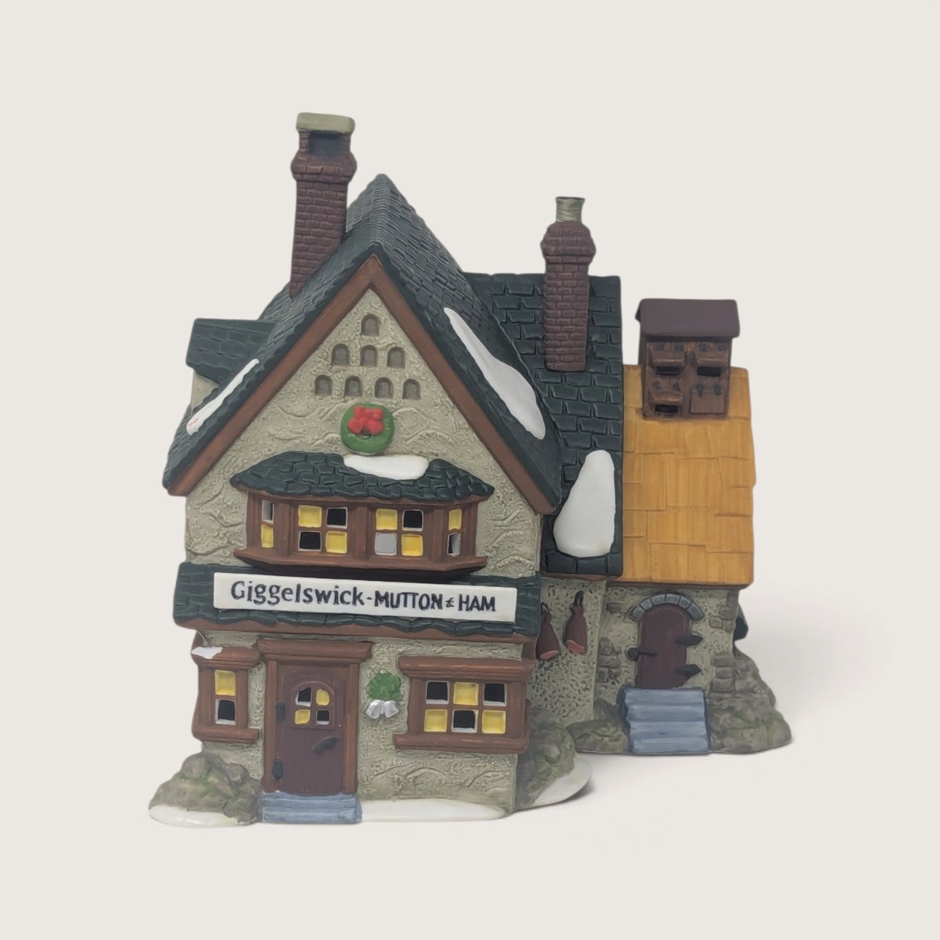 A vintage Department 56 Giggleswick Mutton & Ham lighted ceramic shop building featuring Tudor architecture, a snow-covered roof, and illuminated windows.