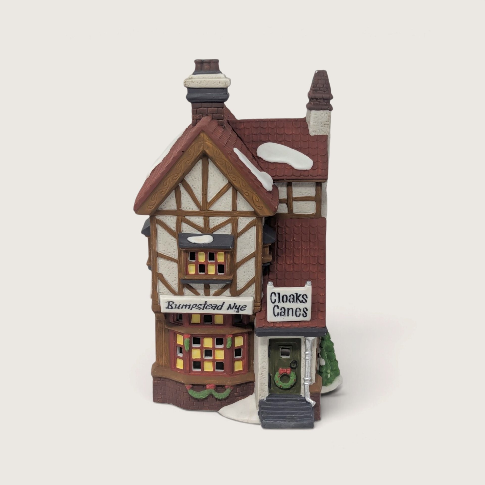 A vintage Department 56 Dickens Village Bumpstead Nye Cloaks and Canes lighted ceramic building featuring Tudor timbering, red shingle roof, and snow accents.