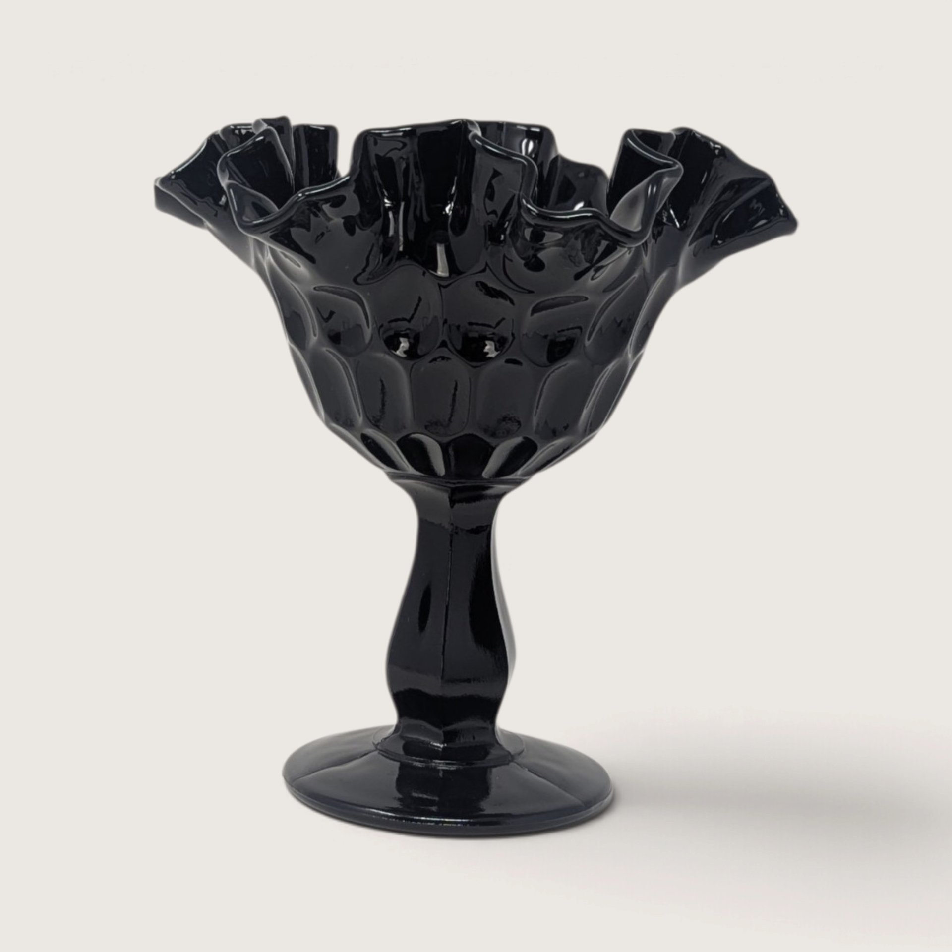 A vintage Fenton black glass compote with an optic thumbprint pattern and a ruffled rim on a neutral background.