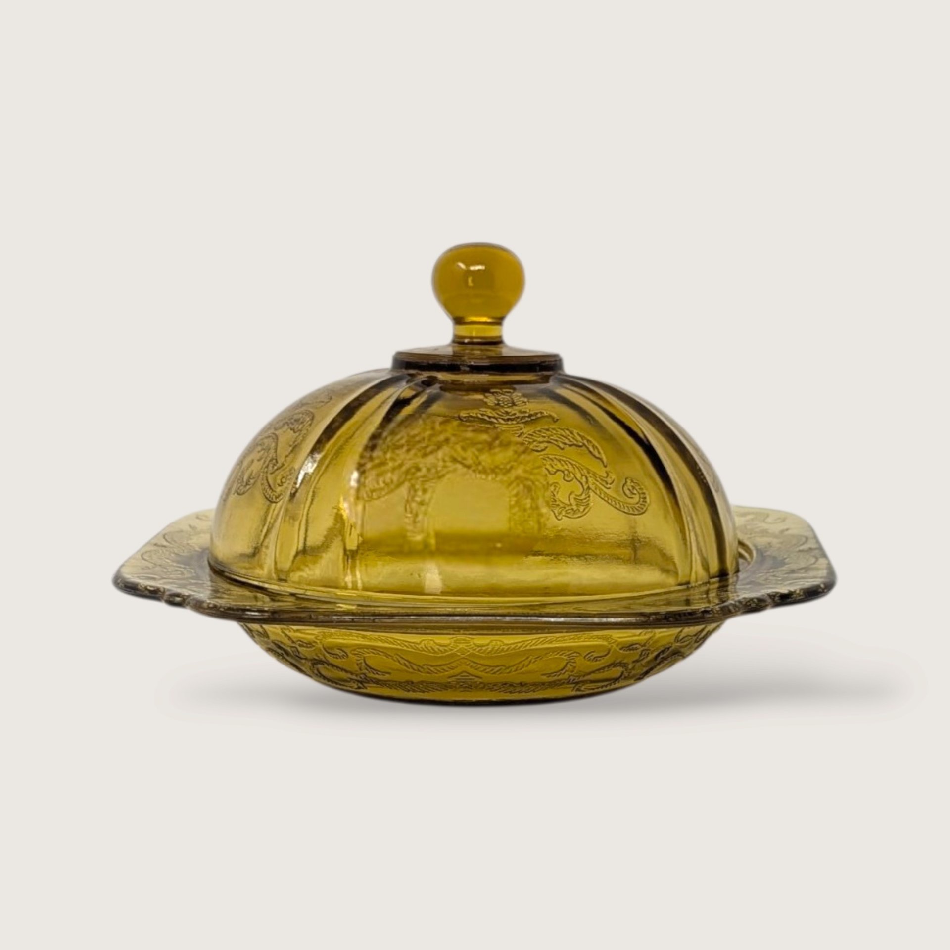 A vintage yellow glass covered butter dish featuring an embossed scroll pattern and domed lid on a neutral background.