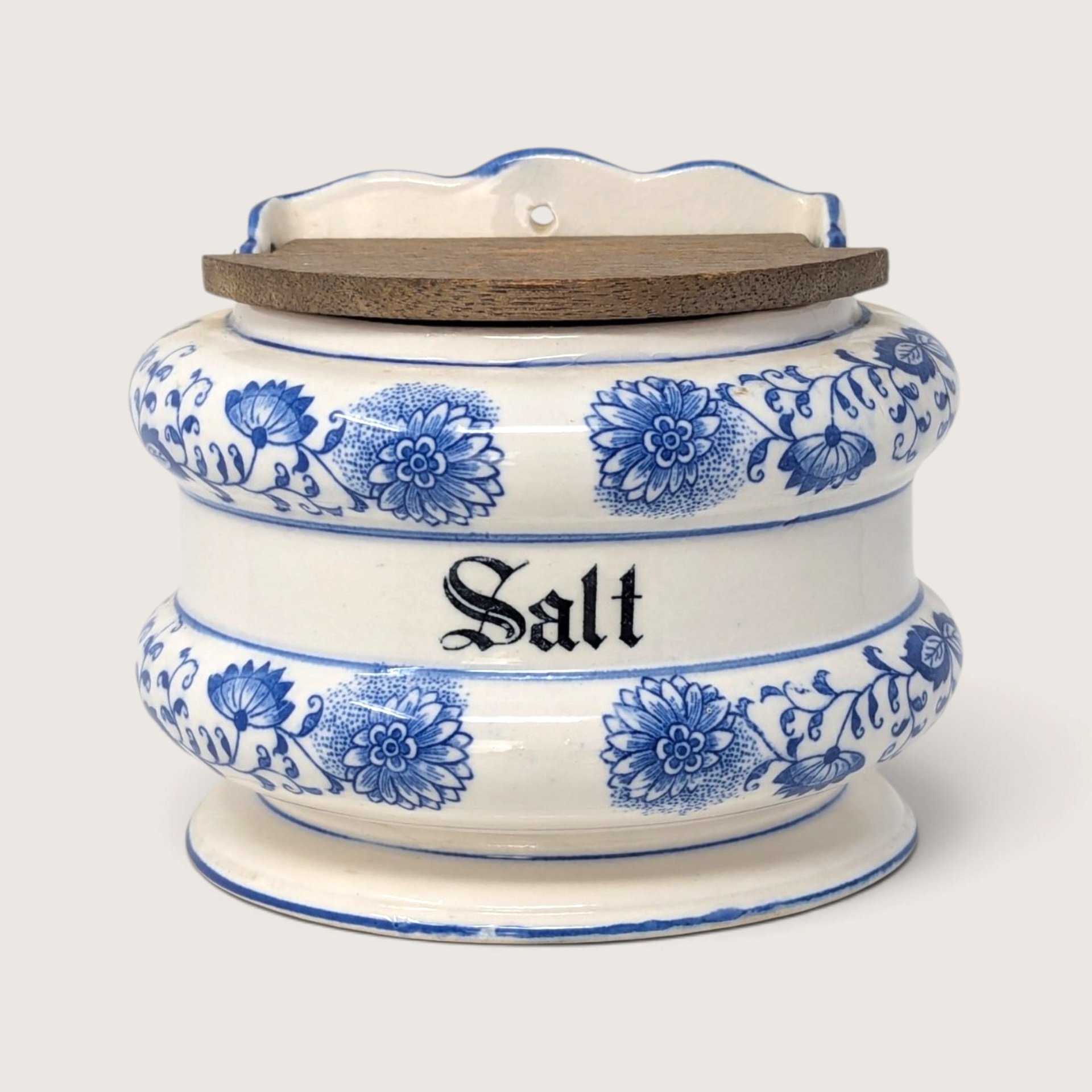 A vintage blue and white ceramic wall-hanging salt bin with a weathered wooden lid and Blue Onion patterns against a neutral background.