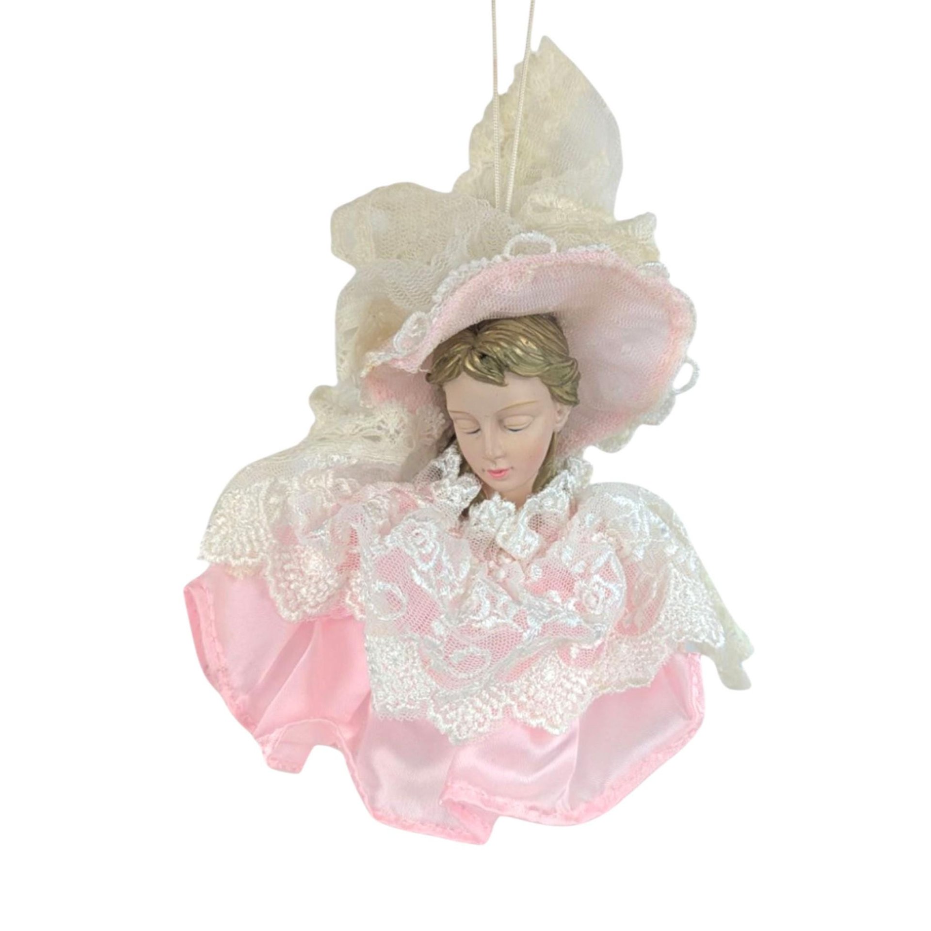 A decorative Christmas ornament featuring a doll head with closed eyes and blonde hair. She is wearing an oversized pink and white lace sun hat and a matching voluminous pink satin dress with a tiered white lace collar and overlay.