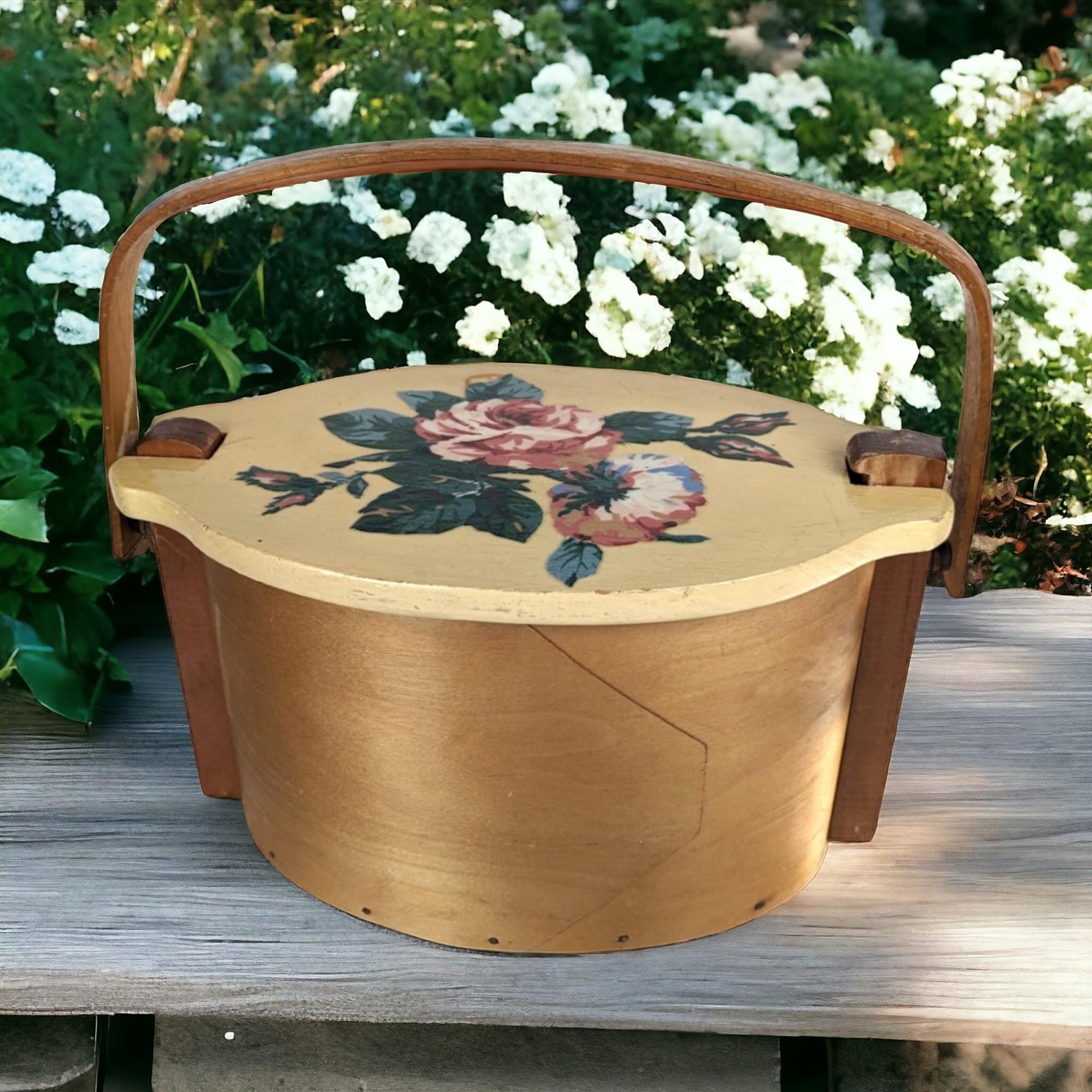 A vintage wooden woven basket with a hinged lid featuring a hand-painted floral design of a pink rose and blue flowers, with a stationary arched wood handle.