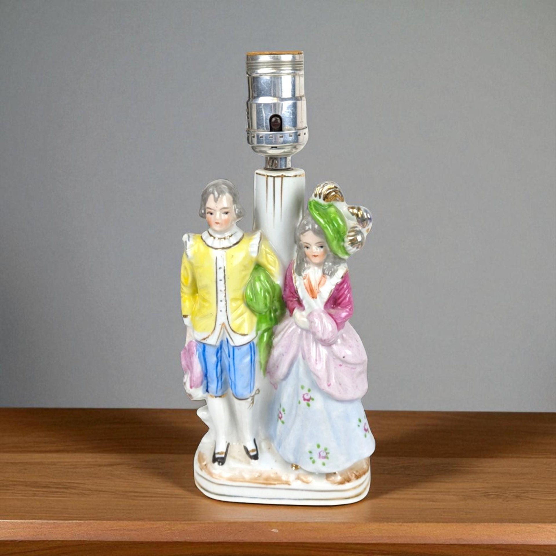 May include: A vintage porcelain lamp featuring a man and woman in period attire. The figures are painted in yellow, blue, pink, and green. The lamp has a silver bulb socket and a white base. The lamp is on a wooden surface.