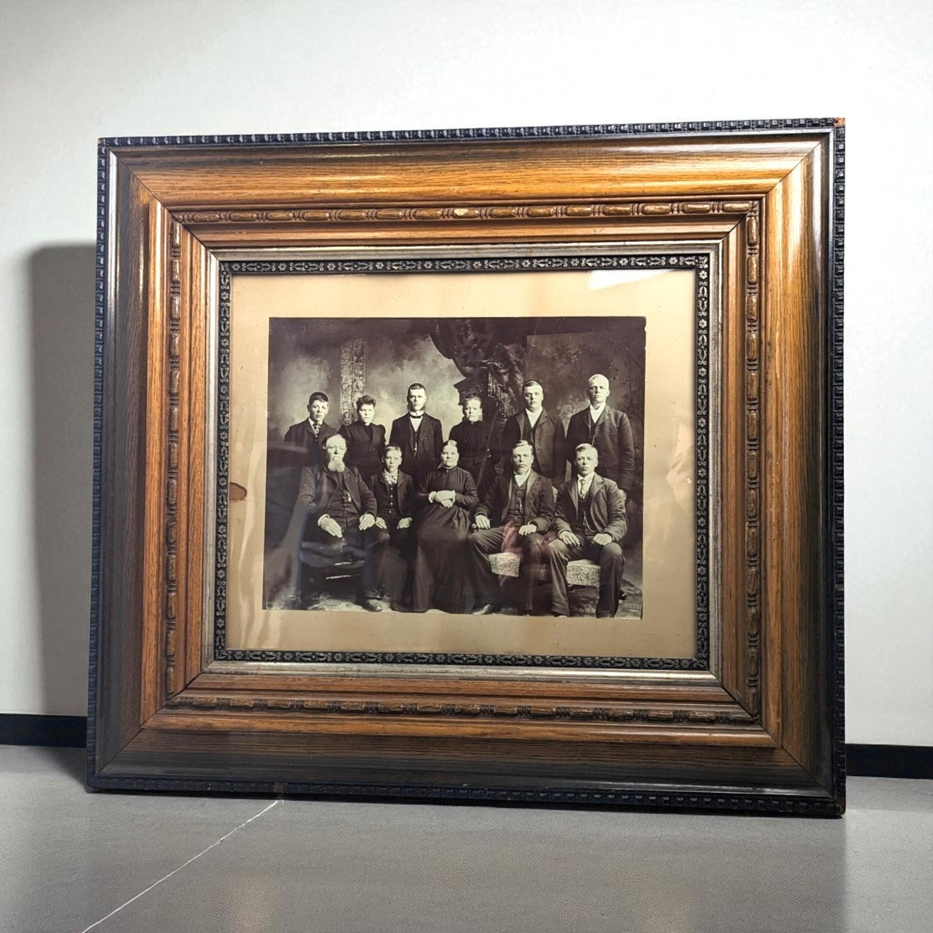 Antique black and white family portrait in a wide, ornate wooden frame. The frame features deep wood grain, silver-toned inner borders, and decorative beadwork. Sepia-toned image shows eleven family members in formal period attire.