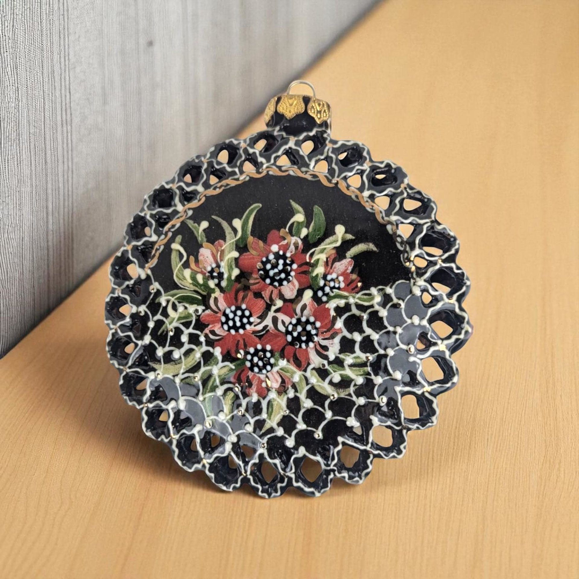 A circular, flat-backed glass ornament featuring a black base with hand-painted red and white floral details. The perimeter is defined by an intricate, die-cut lattice border, while the center displays a bouquet of flowers with white dot accents.