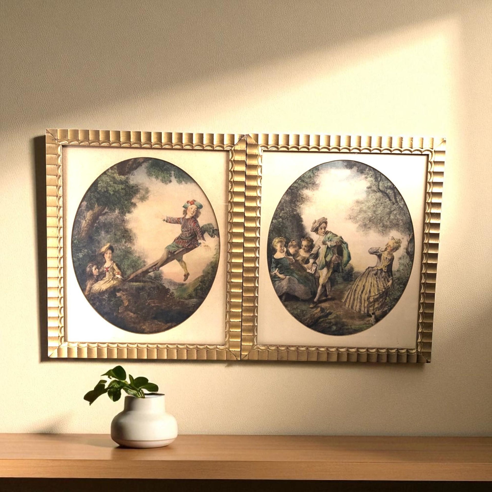 A pair of vintage framed prints depicting 18th-century French Rococo scenes with figures in elegant attire playing on a seesaw and in a park, set in matching fluted gold frames.