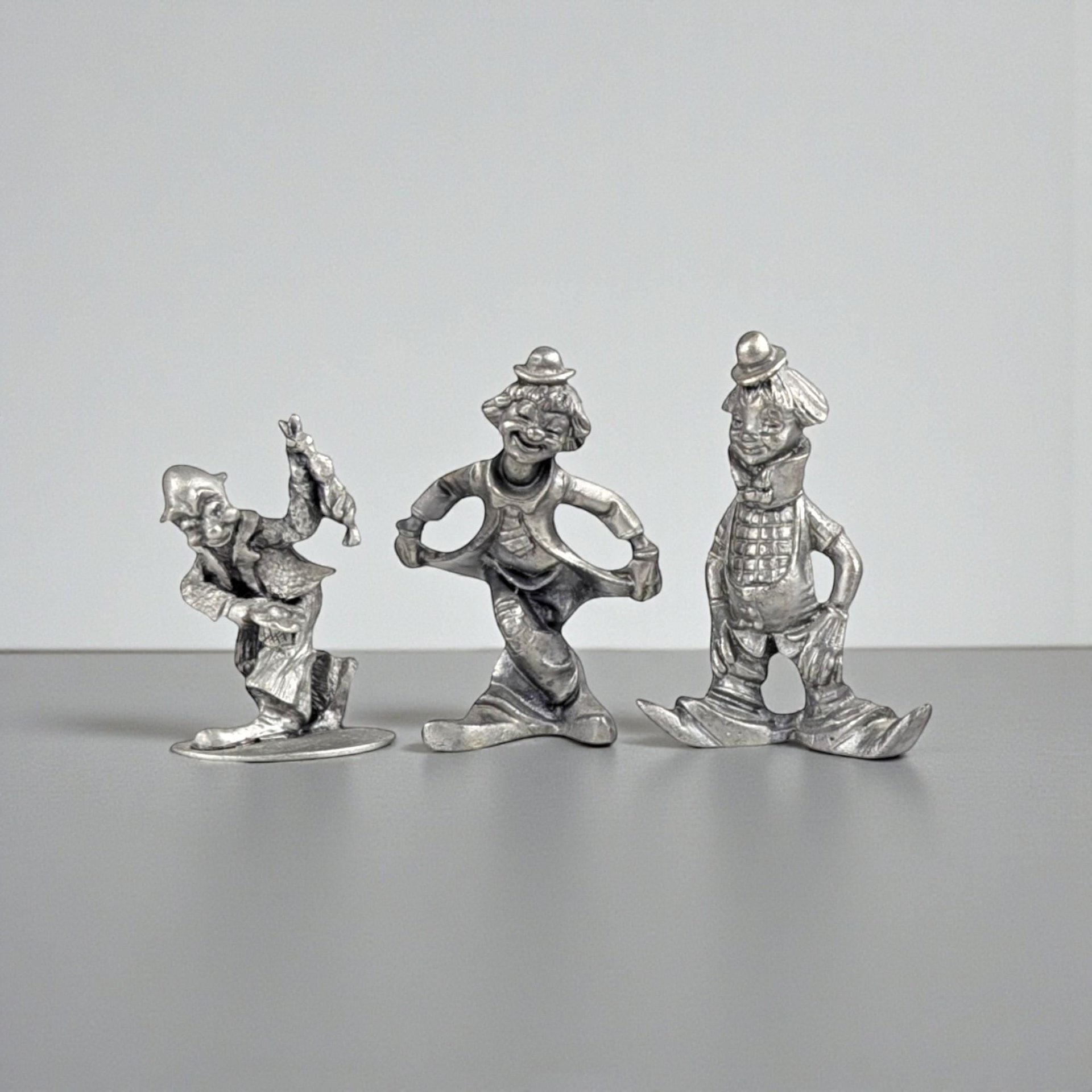 Three small solid pewter clown figurines in various playful poses. The metal has a cool grey, matte patina with dark oxidation in the recessed details. Their silhouettes are slim and whimsical, featuring oversized shoes and tiny hats.