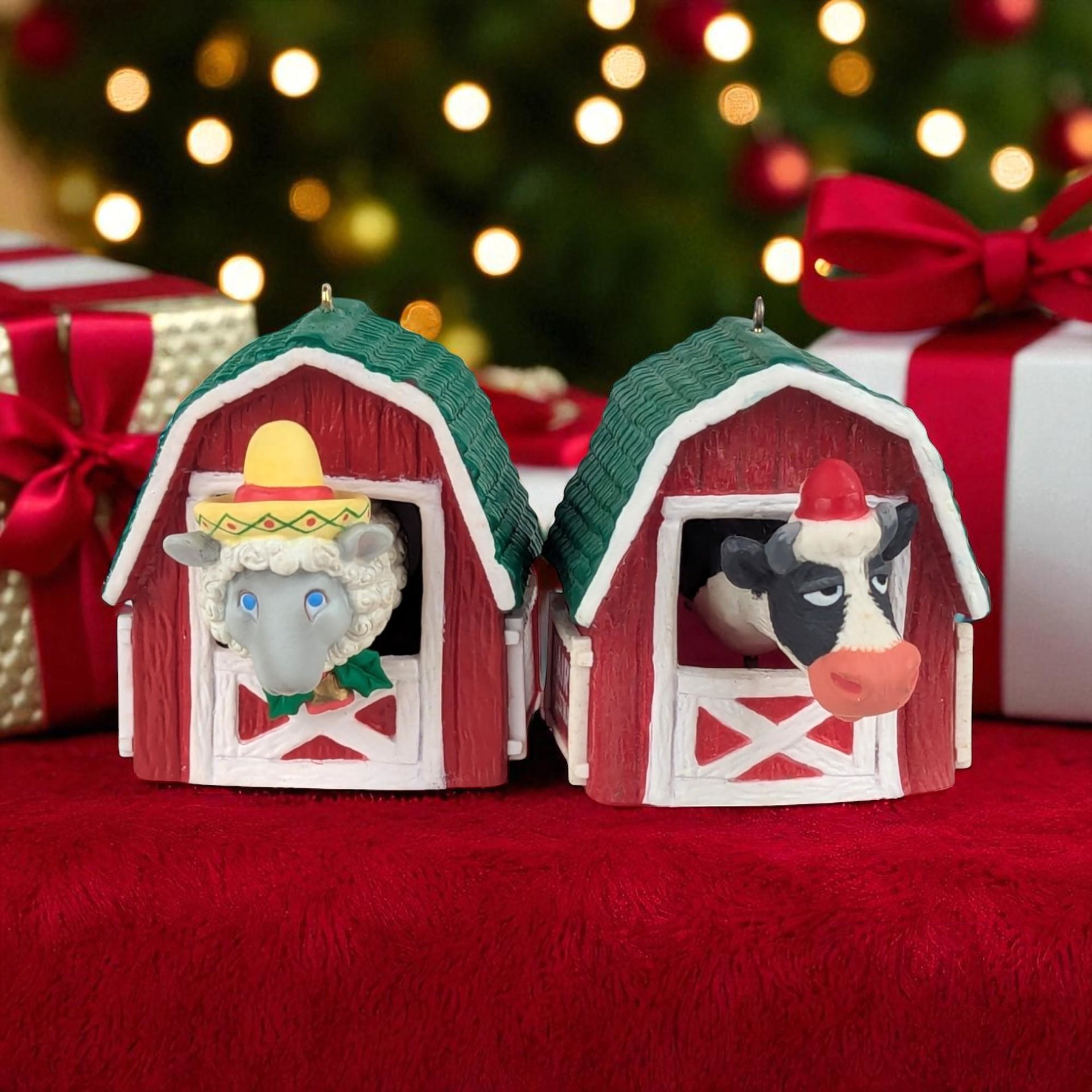 Two vintage Christmas ornaments shaped like small red barns with green roofs; one barn features a black and white cow wearing a red Santa hat, and the other features a white sheep wearing a yellow and red sombrero.