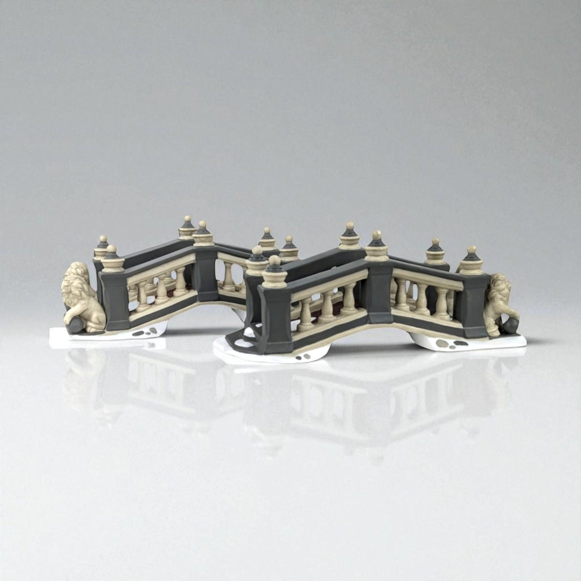 Two miniature porcelain bridge sections with a matte charcoal and cream finish. Each bridge features arched balustrades, decorative finials, and a sculpted lion seated at the base. The snowy white foundations suggest a winter village setting.