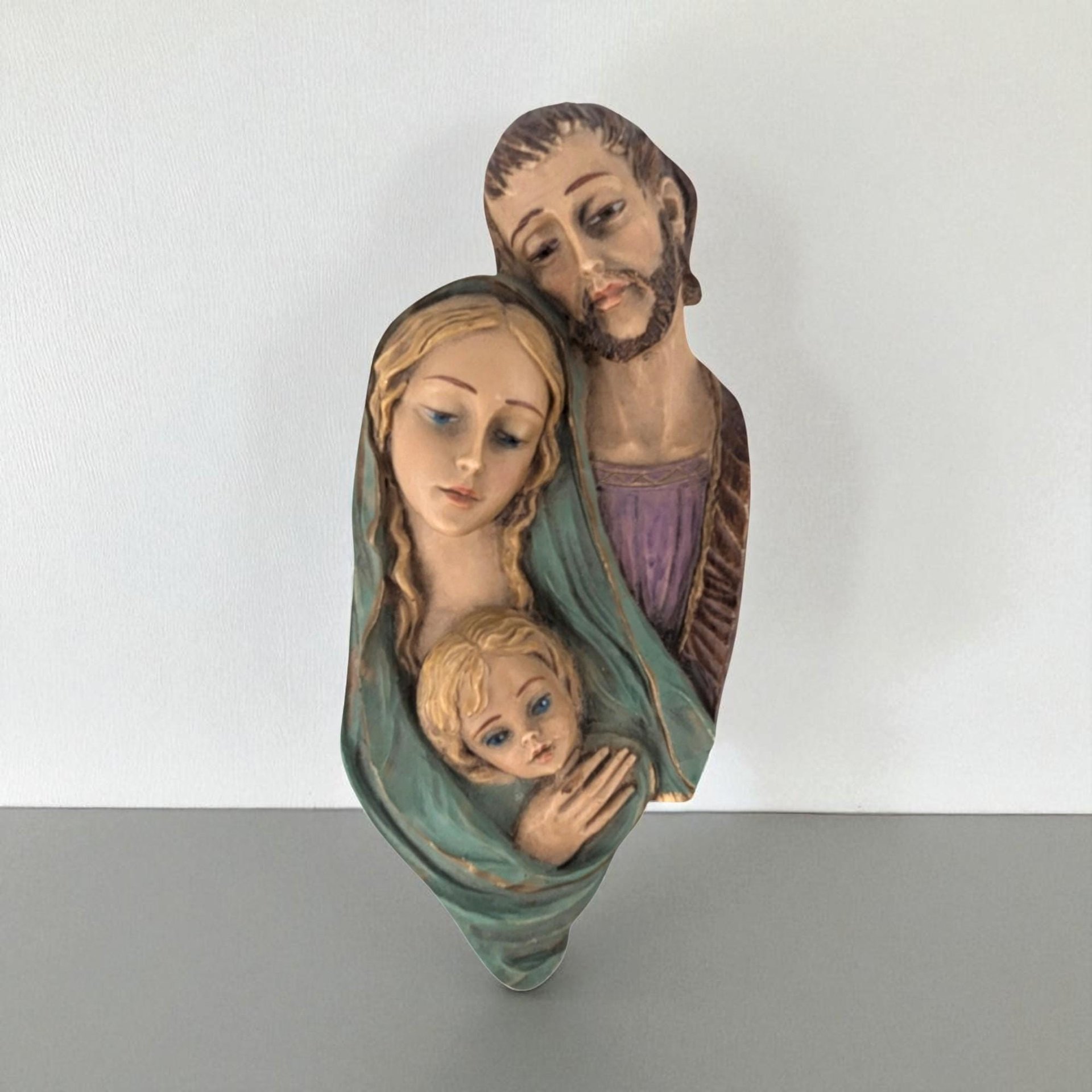 A high-relief wall plaque depicting the Holy Family. The figures feature a matte, hand-painted finish in muted tones of teal, violet, and earth browns. The sculpture highlights smooth facial features, flowing drapery, and a vertical, tapering silhouette.