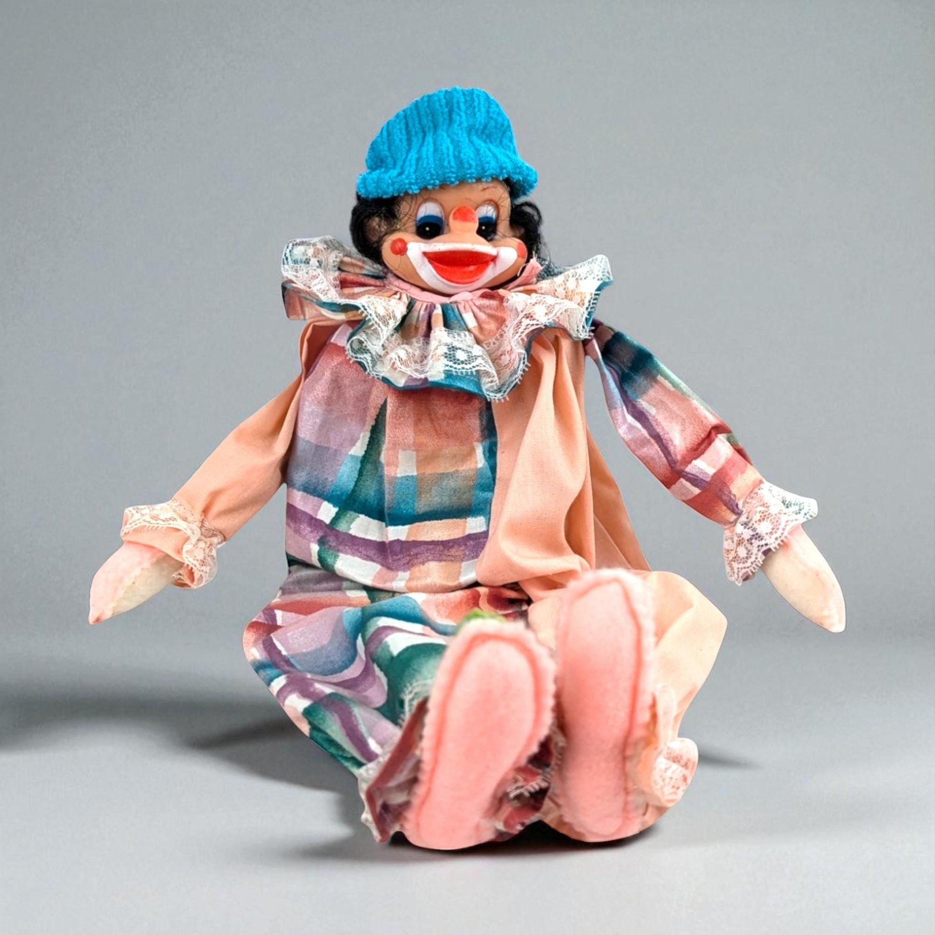 A vintage clown doll with a molded plastic face, a bright blue knit hat, and a ruffled lace collar. The body features a muted pastel plaid pattern in peach, teal, and purple. The hands and feet are soft peach fabric.