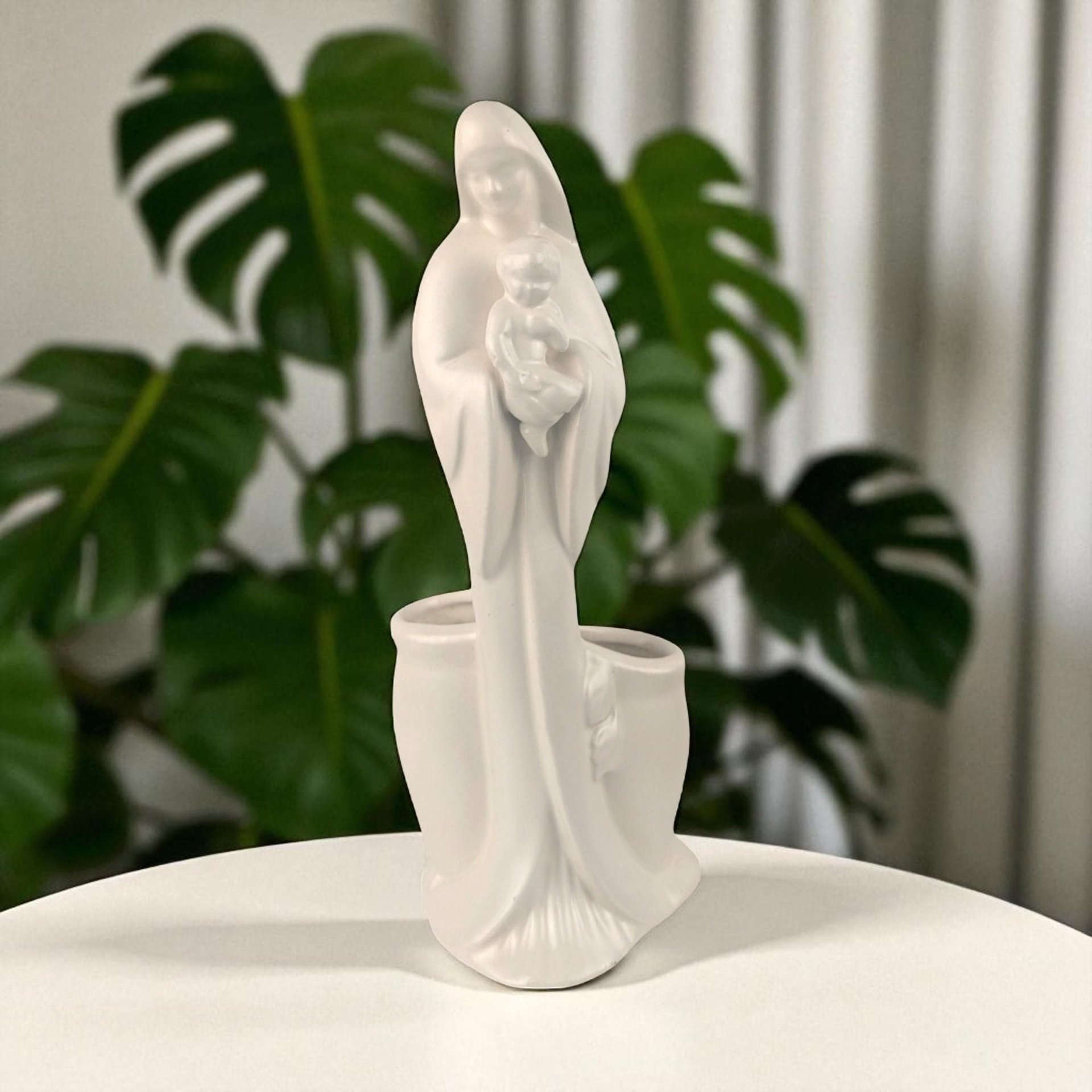 A matte white ceramic figurine of the Madonna holding the infant Jesus. The texture is smooth and non-reflective. The silhouette is tall and fluid, featuring a hollow planter well integrated into the back of the figure.