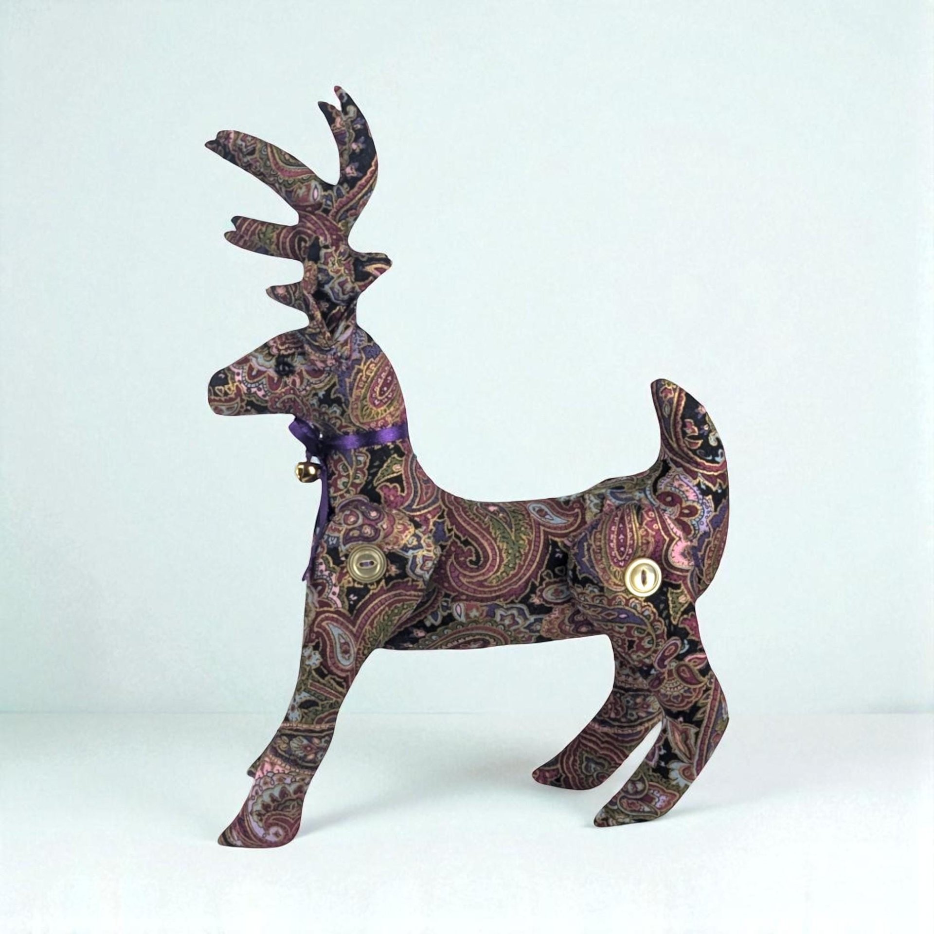 A standing plush reindeer made from fabric with an intricate purple and earth-toned paisley pattern. It features large antlers, button-jointed legs, and a purple ribbon with a small gold bell around its neck.