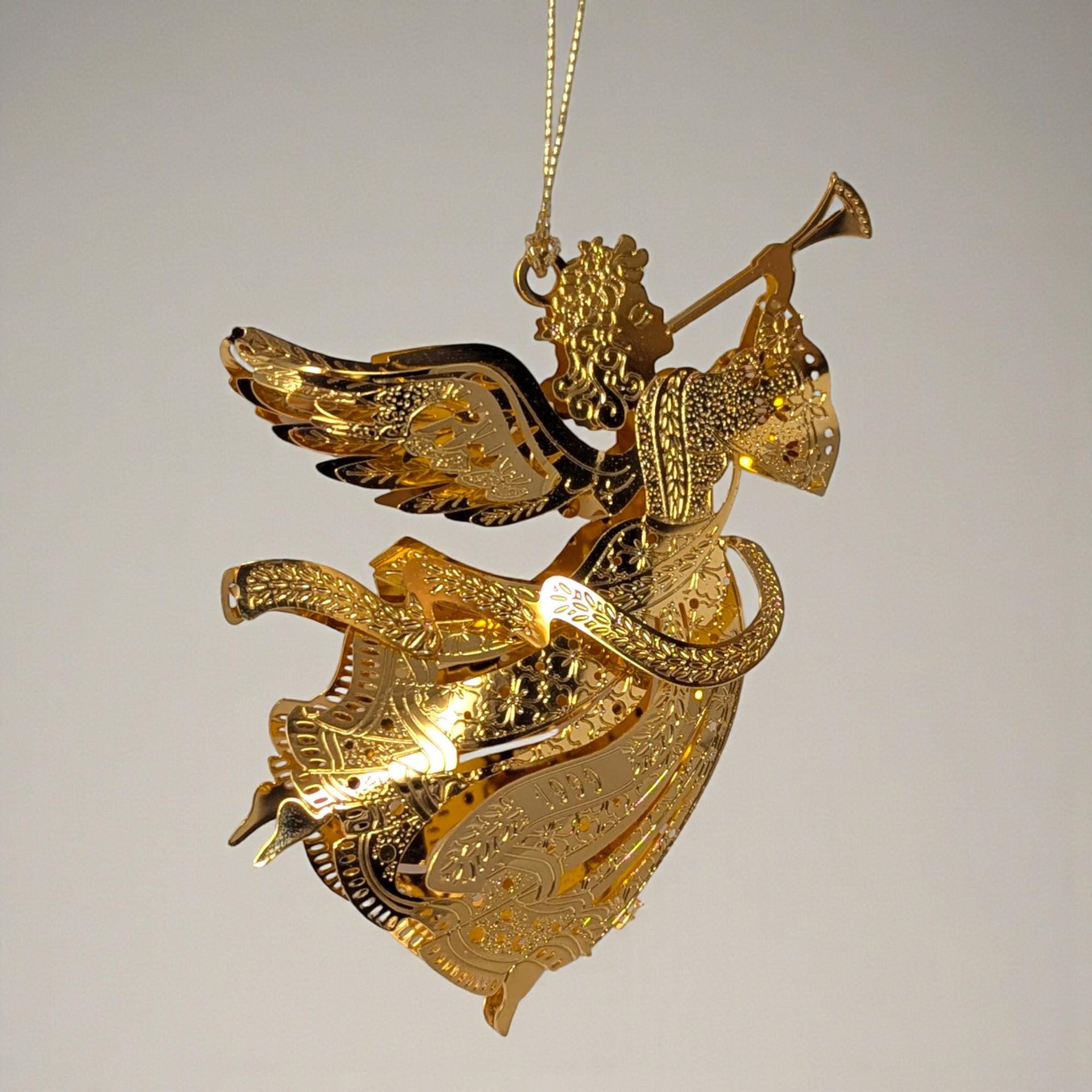 Polished gold-tone metal angel ornament with an openwork silhouette. The angel has feathered wings, holds a small star, and is framed by an elegant scrollwork border featuring the year 1999.