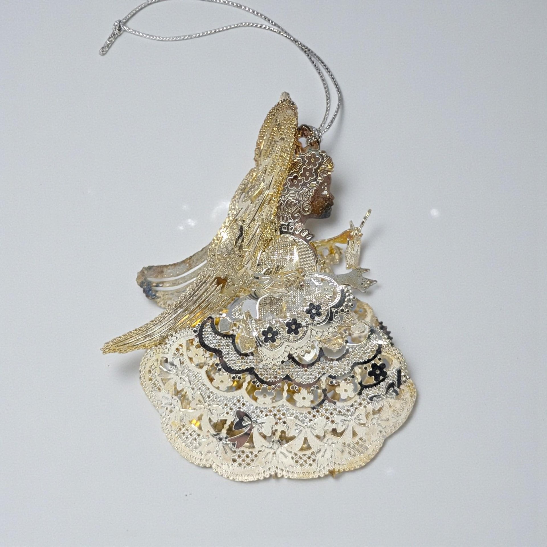 Silver plated metal ornament of a praying angel with large feathered wings and a long flowing gown. The surface has a soft aged patina with visible tarnish in the recessed details.