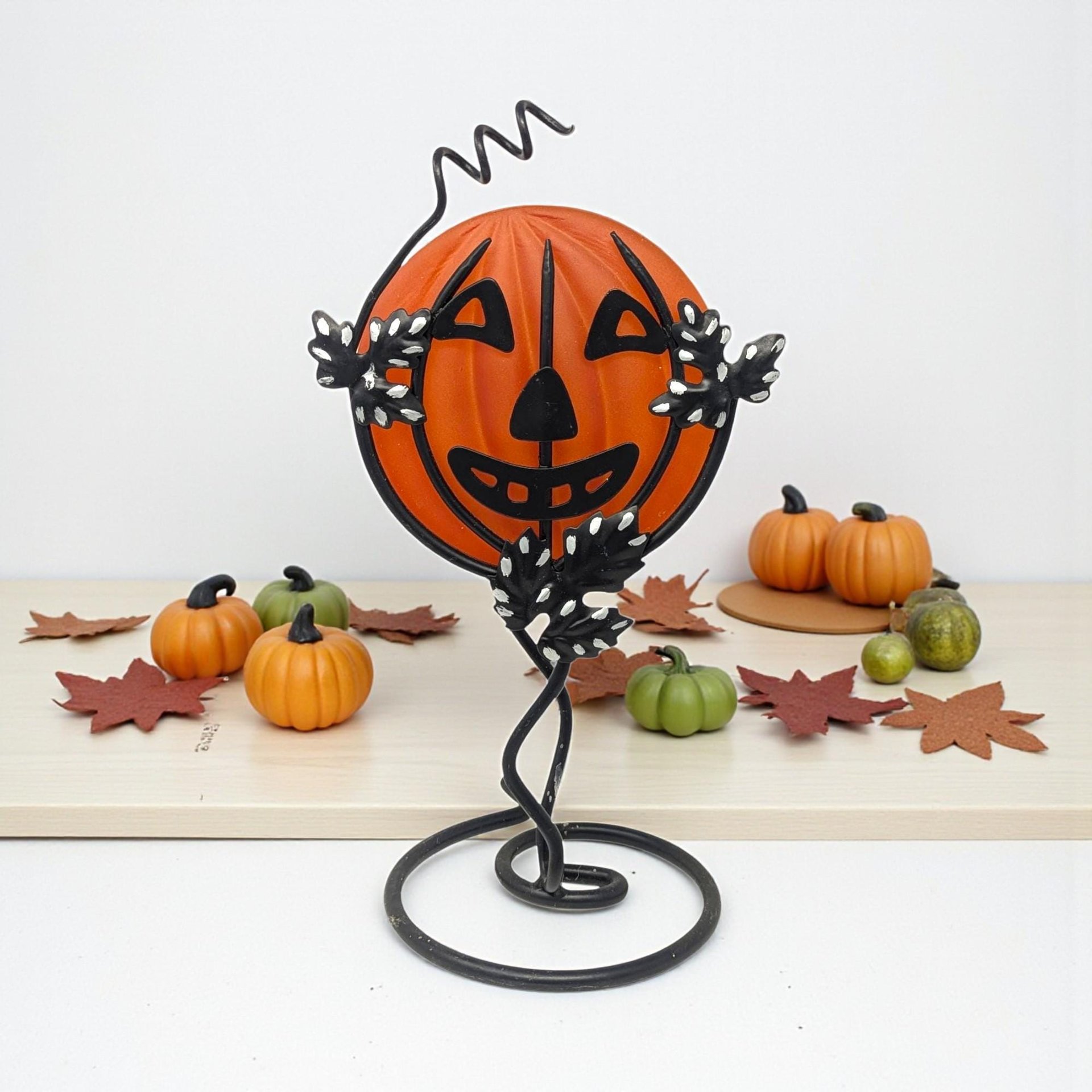 A frosted orange glass pumpkin face is cradled within a black wrought iron stand. The metal frame features a coiled base, climbing vine-like tendrils, and speckled leaves. A whimsical wire curl extends from the top of the jack-o'-lantern silhouette.