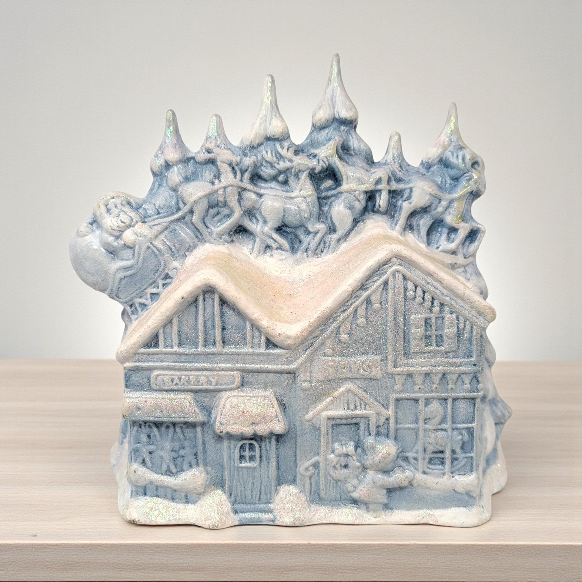 A wide ceramic Christmas figurine featuring a hand-painted winter scene. The piece shows a high-gloss glaze with vibrant red and green holiday tones, depicting festive characters in a traditional mid-century folk art style.