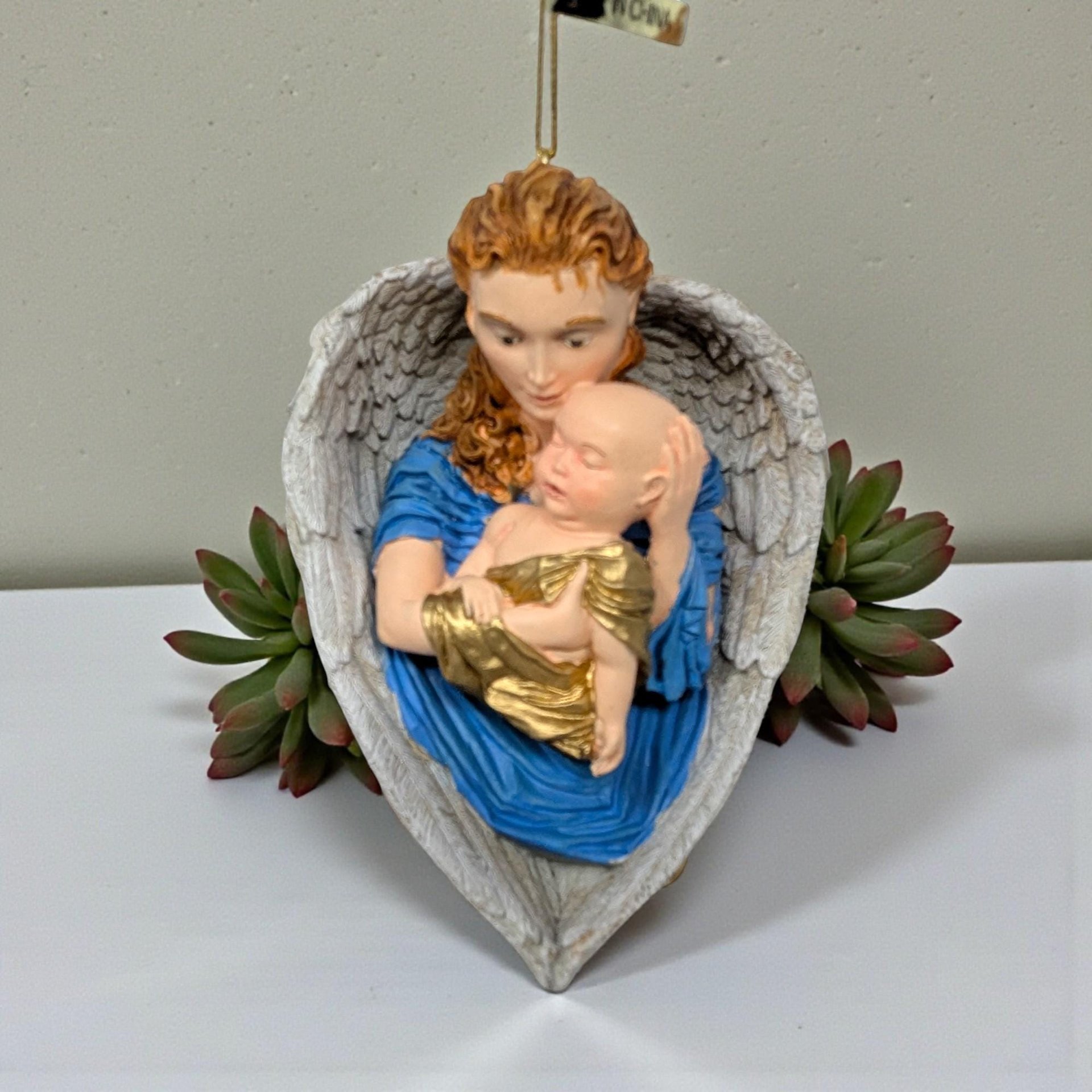 A sculpted resin ornament featuring a red-haired angel in a vibrant blue robe cradling a baby in gold swaddling. The figures are framed by large, textured white wings that form a heart shape at the base.