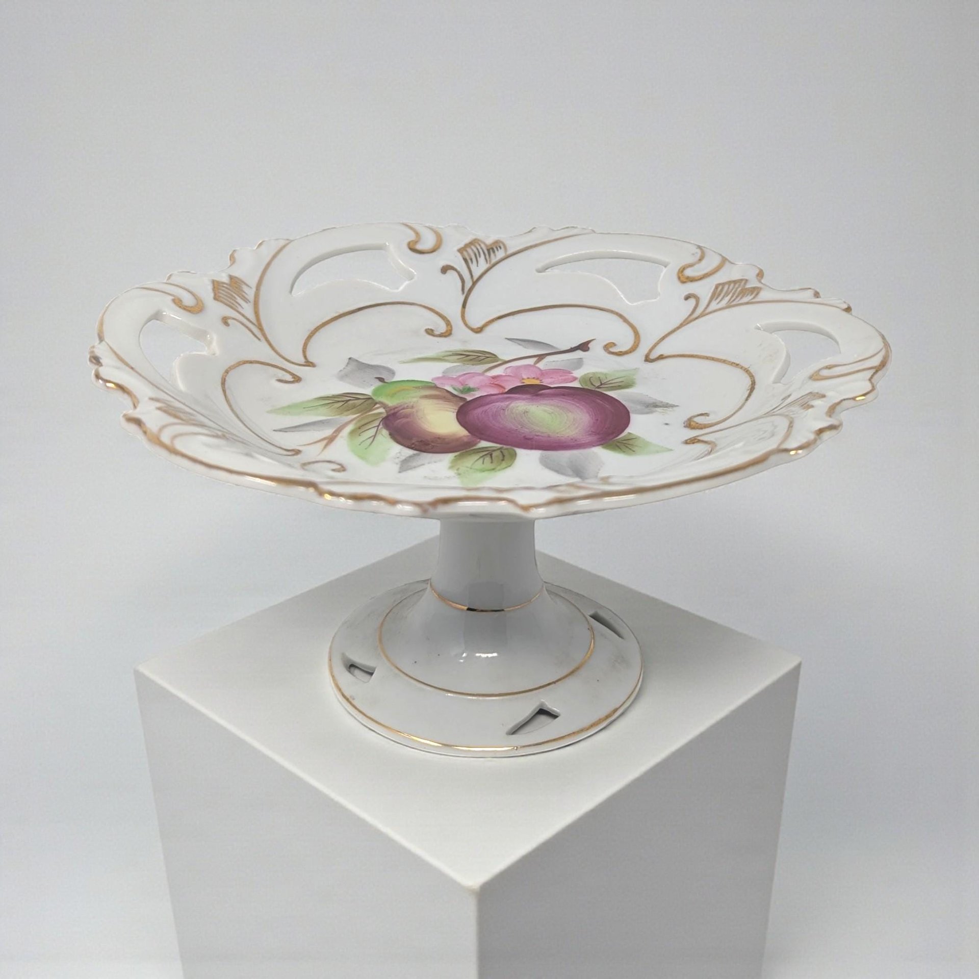 A white porcelain pedestal dish featuring a hand-painted plum and pear motif at the center. The shallow bowl has a decorative pierced or reticulated rim accented with gold filigree and a matching footed base.
