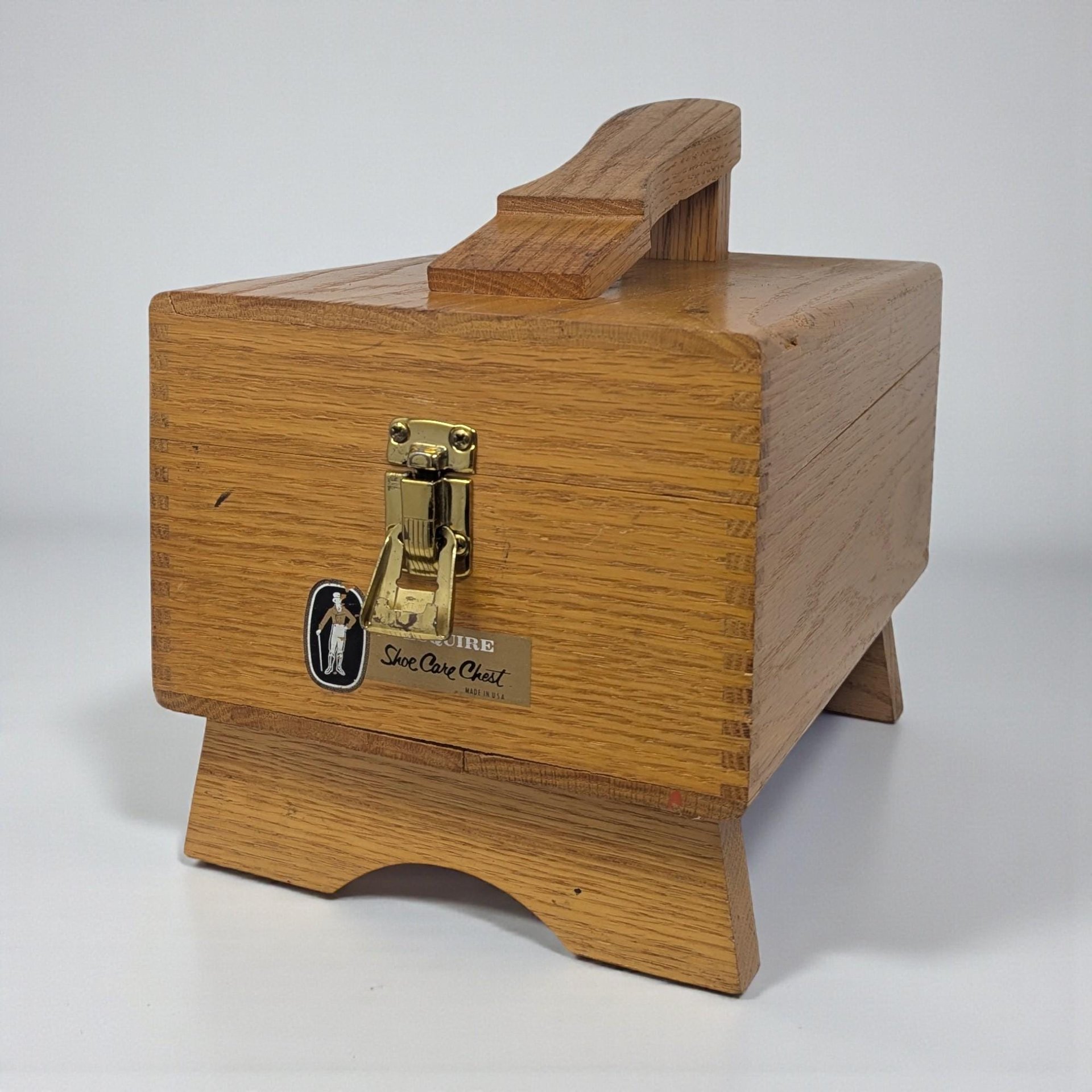 A light-toned oak shoe shine box featuring a built-in footrest on top. The chest has finger-jointed corners, a brass-tone metal latch, and an original Esquire Shoe Care Chest label on the front.
