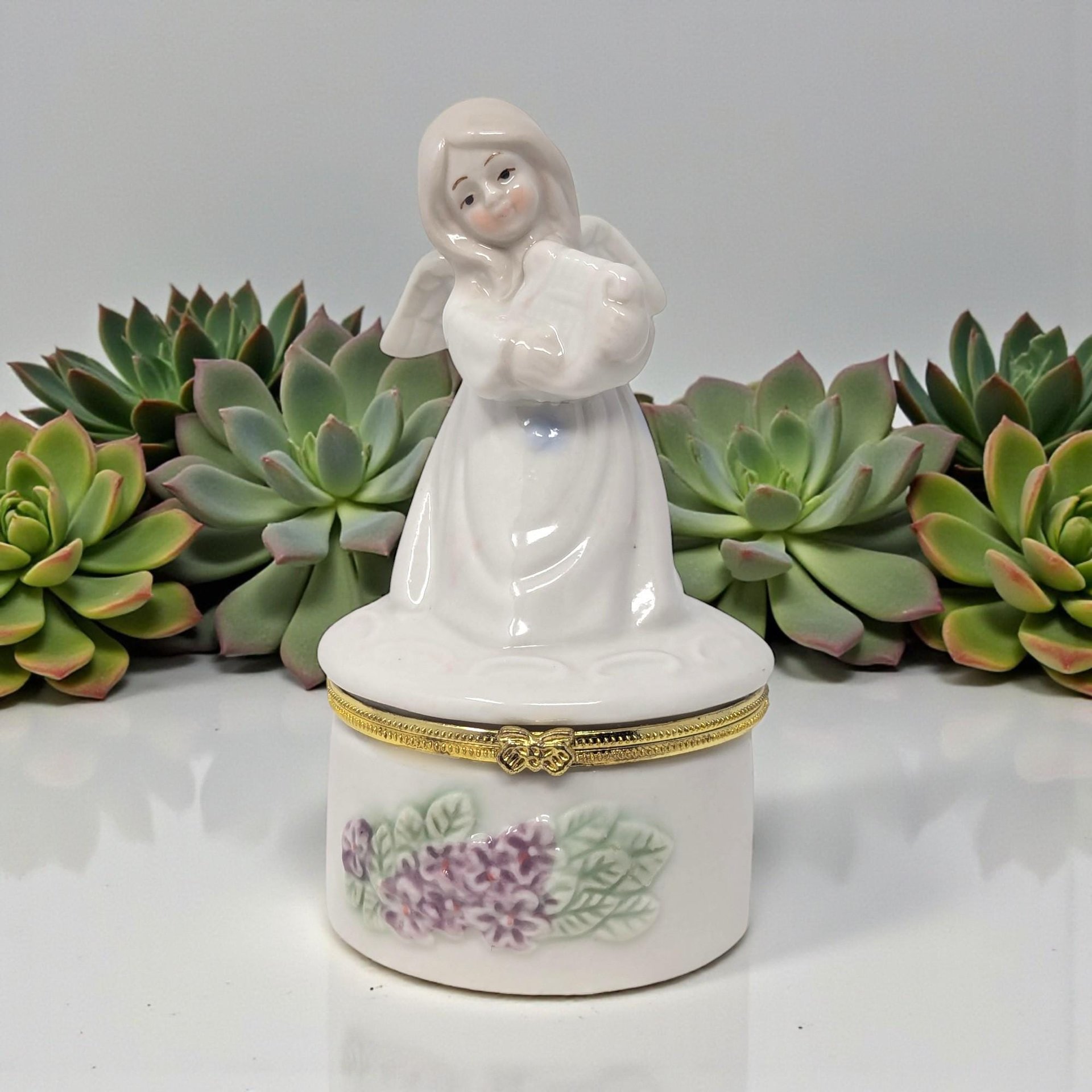 A slender white ceramic figurine of an angel playing a gold-accented harp. The base of her gown is decorated with delicate purple floral sprigs and opens to reveal a small storage compartment.