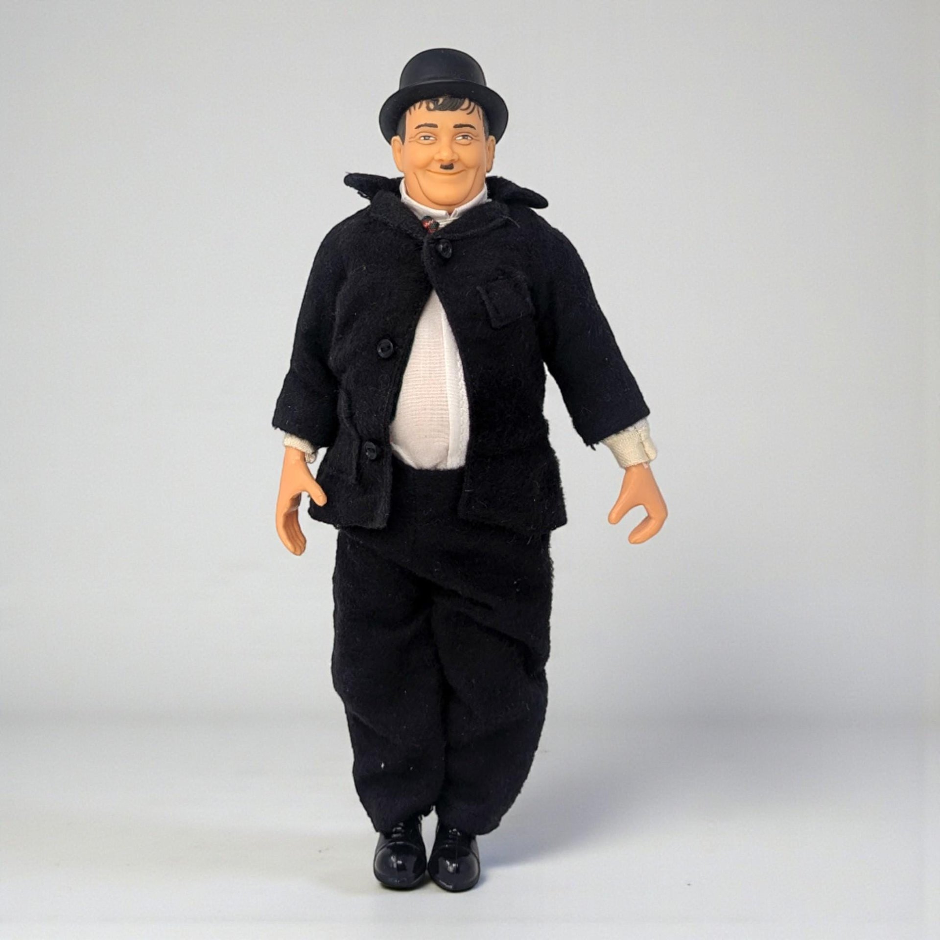 A vinyl character doll of comedian Oliver Hardy featuring his iconic bowler hat, black suit, and mustache. The figure has a rounded physique and visible joints at the elbows and knees.