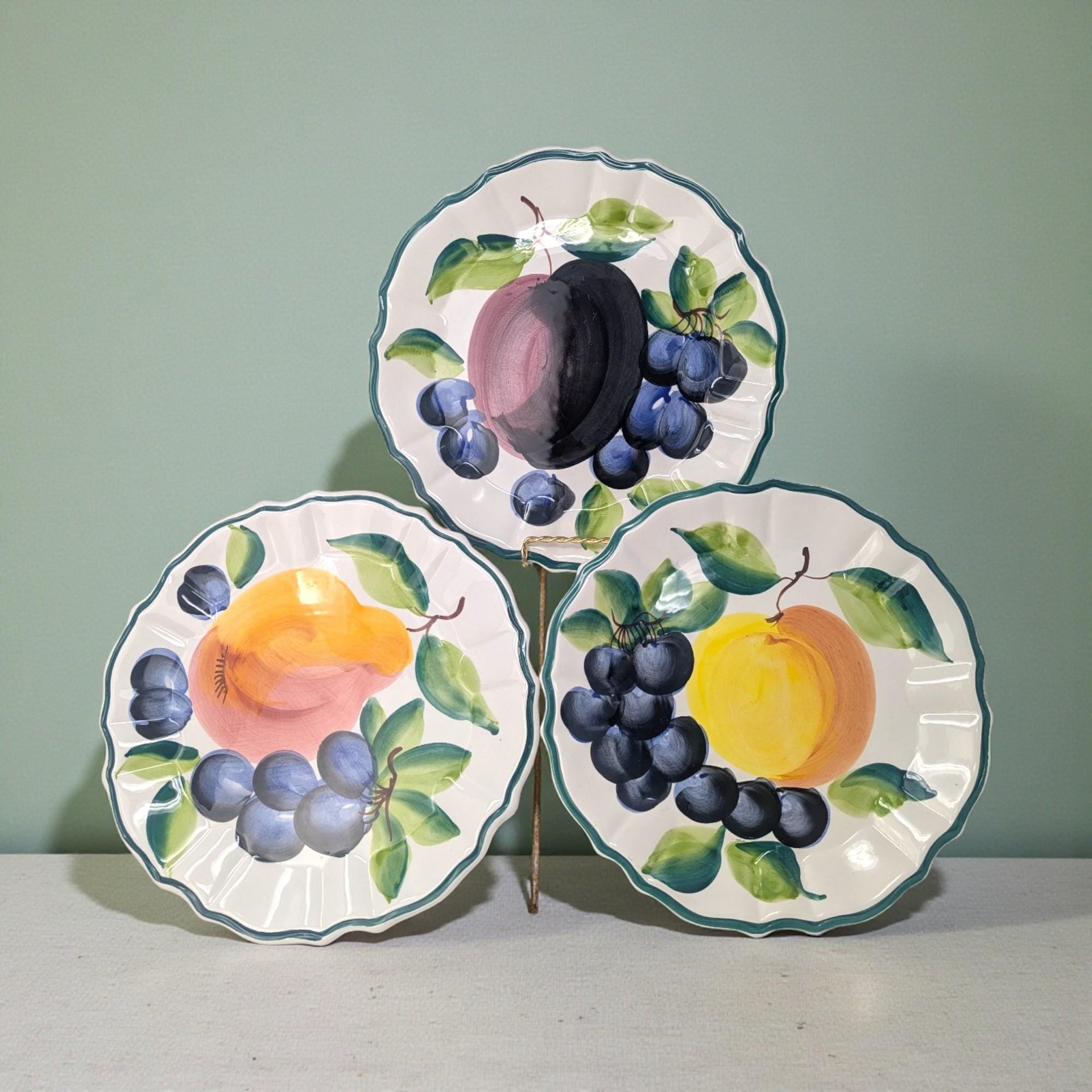 A set of three white ceramic plates with scalloped edges. Each plate features a vibrant, hand-painted fruit motif in the center—grapes, lemons, and plums—surrounded by delicate green leaf accents and a glazed finish.