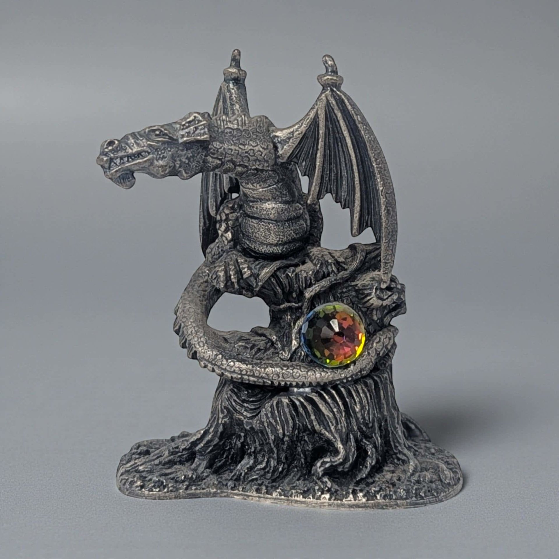 A solid pewter dragon figurine with a cool grey metallic luster. The dragon is perched on a gnarled tree stump with its wings partially unfurled. Fine scales and textured bark detail the surface, featuring a small faceted rainbow crystal orb.