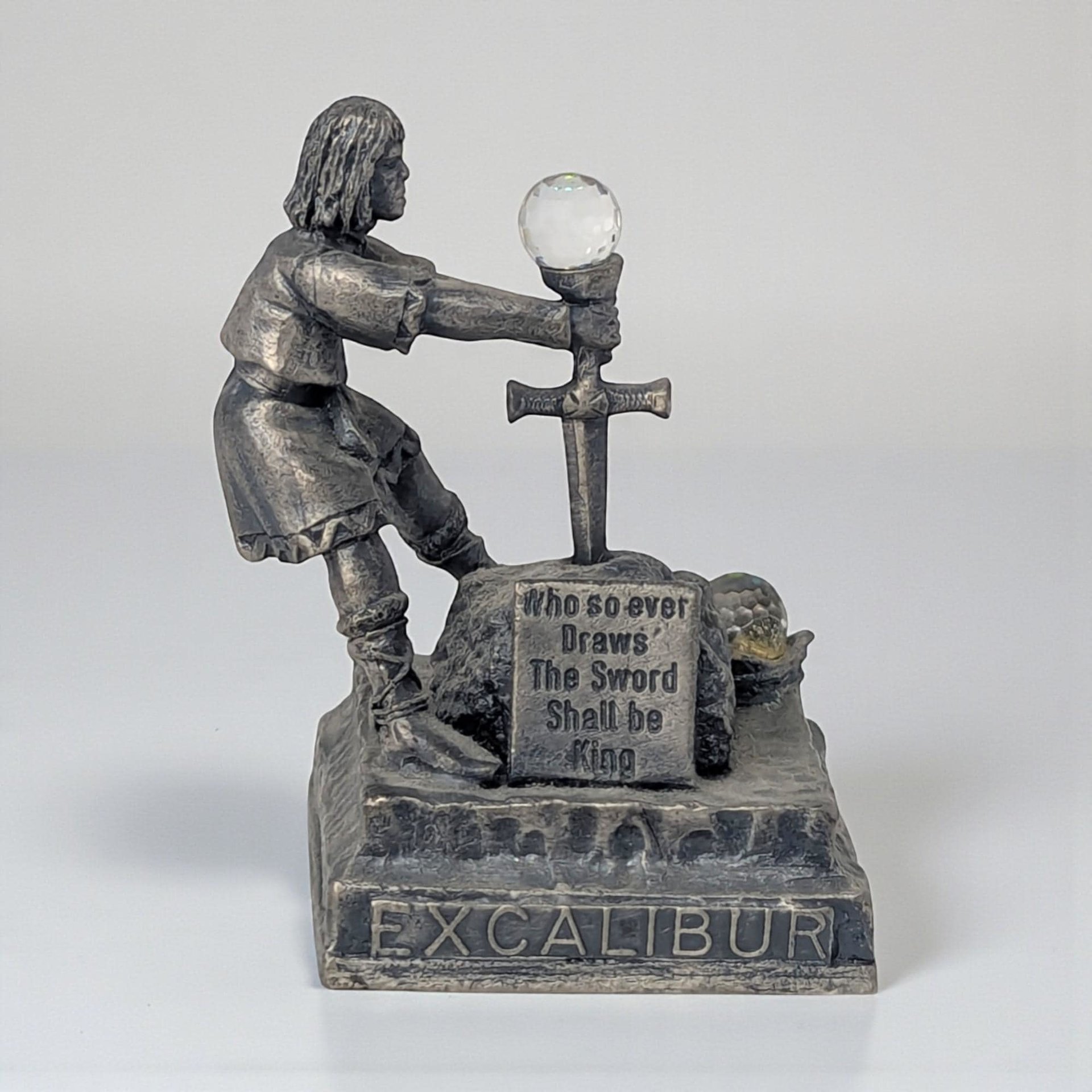 A matte gray pewter figurine of King Arthur pulling the sword Excalibur from a stone. The metal features a weathered, dark patina. A small multifaceted clear crystal sits atop the sword hilt.