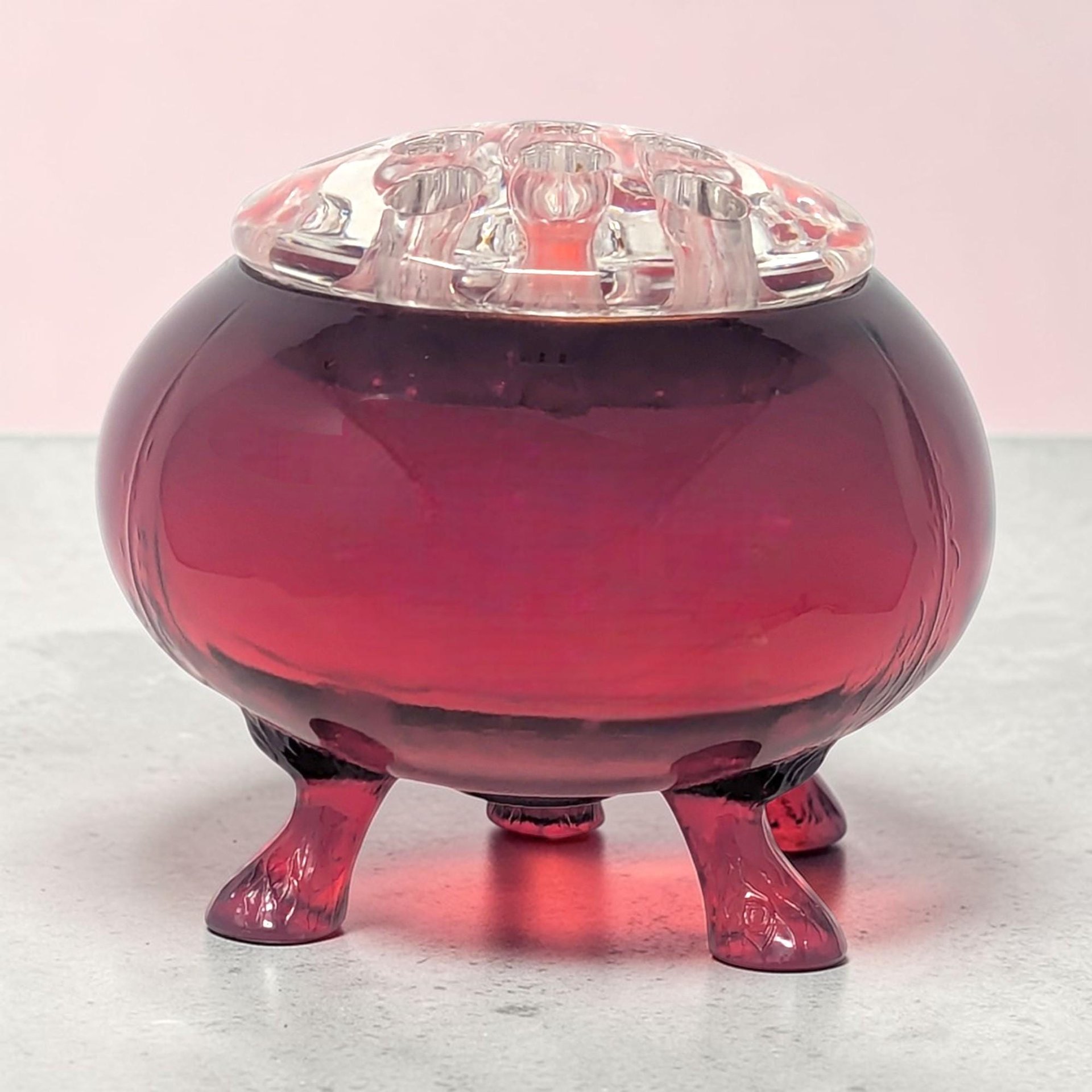 Deep ruby red glass flower frog with a spherical body and flattened top containing multiple holes. Three curled clear glass feet support the vibrant red vessel. High-gloss finish with a rich, saturated glow.
