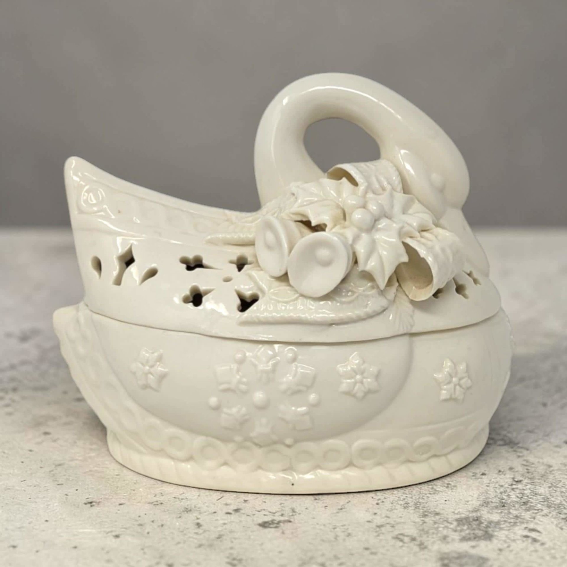 A creamy white ceramic swan-shaped bowl with a matching lid. The lid features decorative holly leaves, berries, and bells tucked under the swan's curved neck. The base is embossed with snowflakes and a braided border.