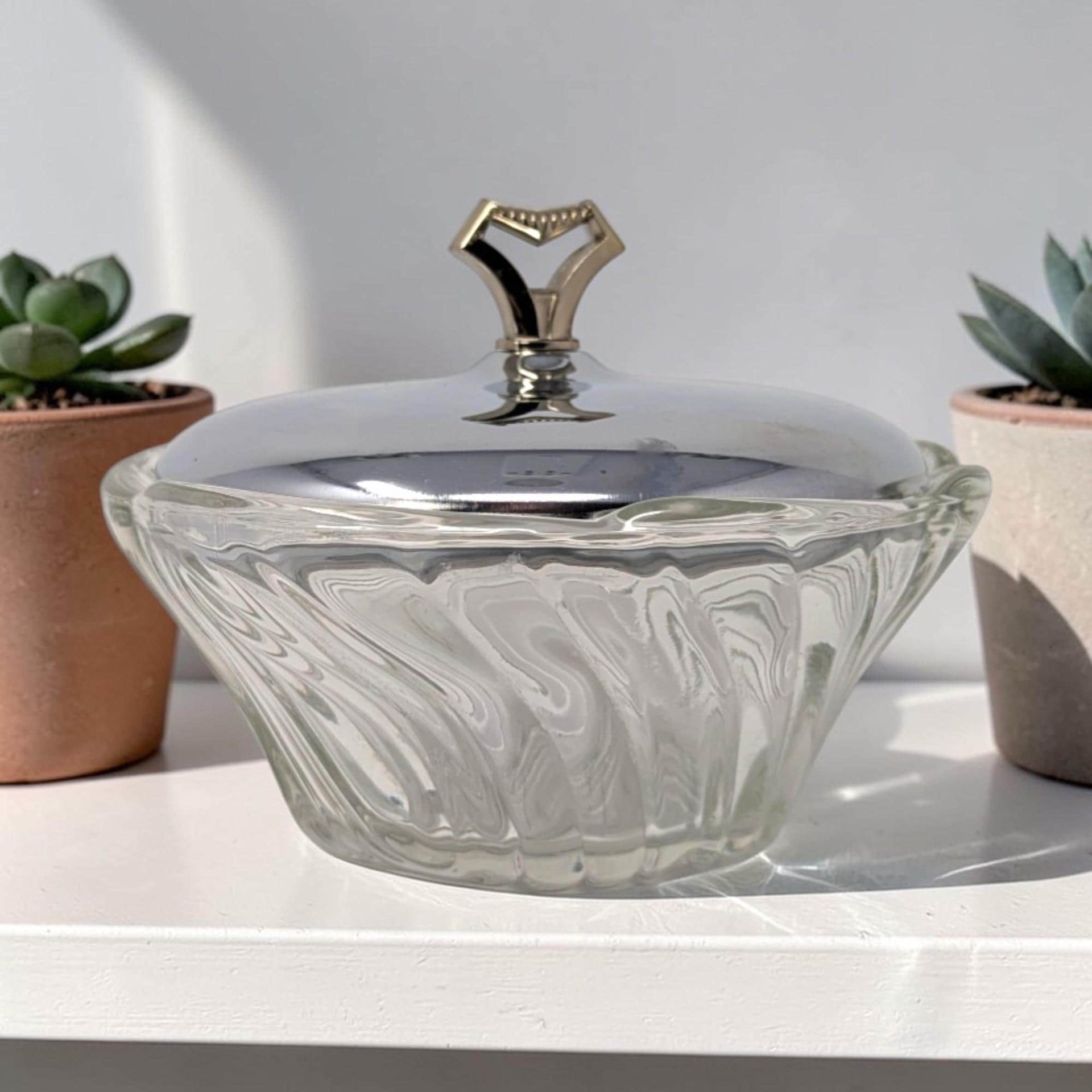 A clear glass bowl with a deep swirled or ruffled texture around the exterior. It is topped with a smooth, reflective silver-toned metal lid featuring a geometric, gold-toned pedestal handle.
