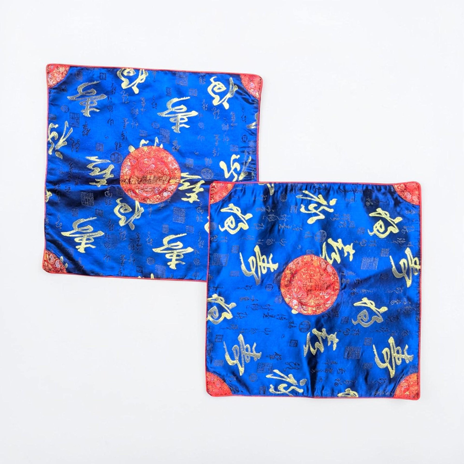 Two square satin pillow covers in a deep cobalt blue with metallic gold calligraphic characters. A central red circular medallion with gold embroidery anchors the design, and red brocade corners finish the edges.