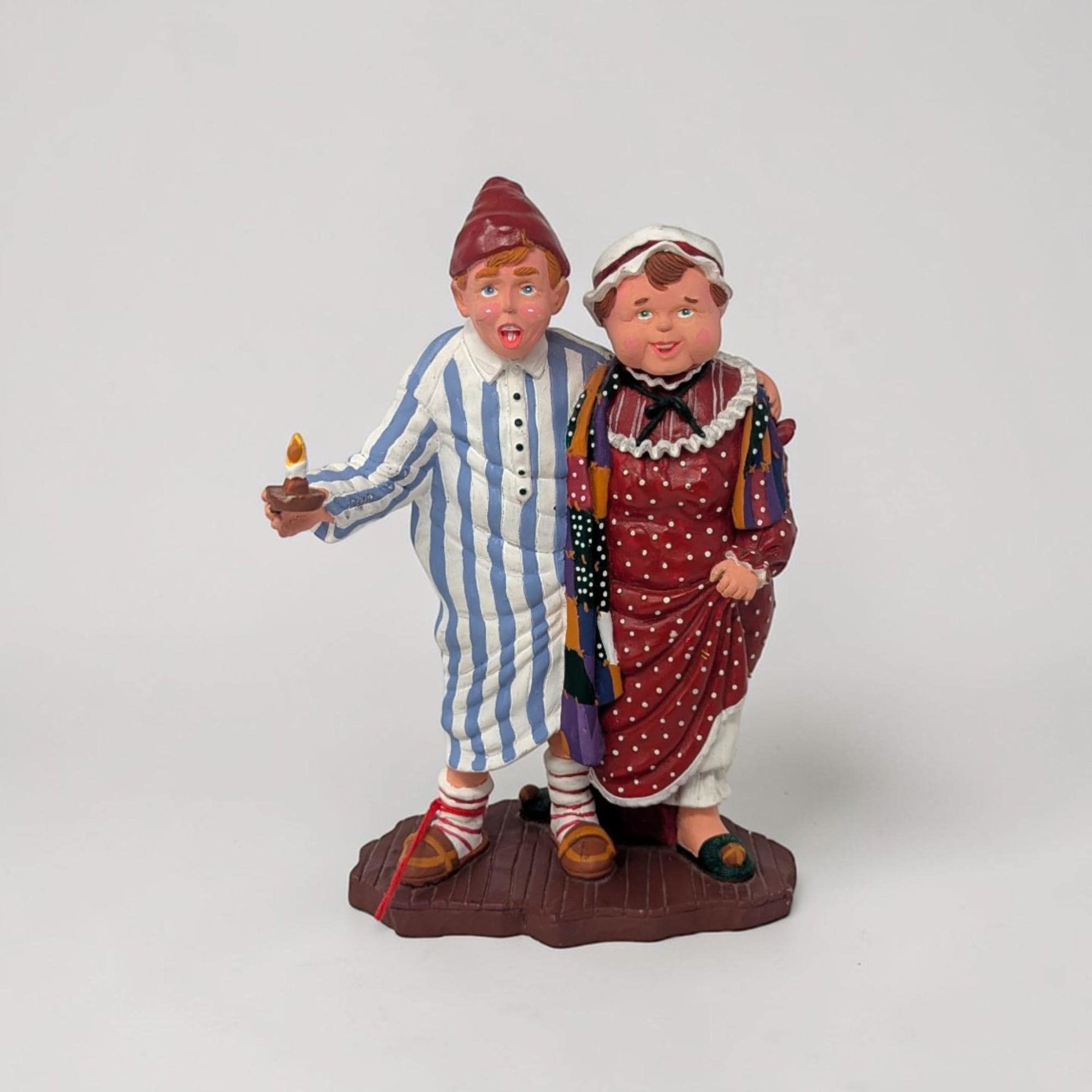 Hand-painted porcelain figurine of a man and woman in Victorian nightwear. "Mama" wears a white kerchief and pink robe, while "Papa" is in a blue striped nightshirt and red cap. They stand together on a brown base.