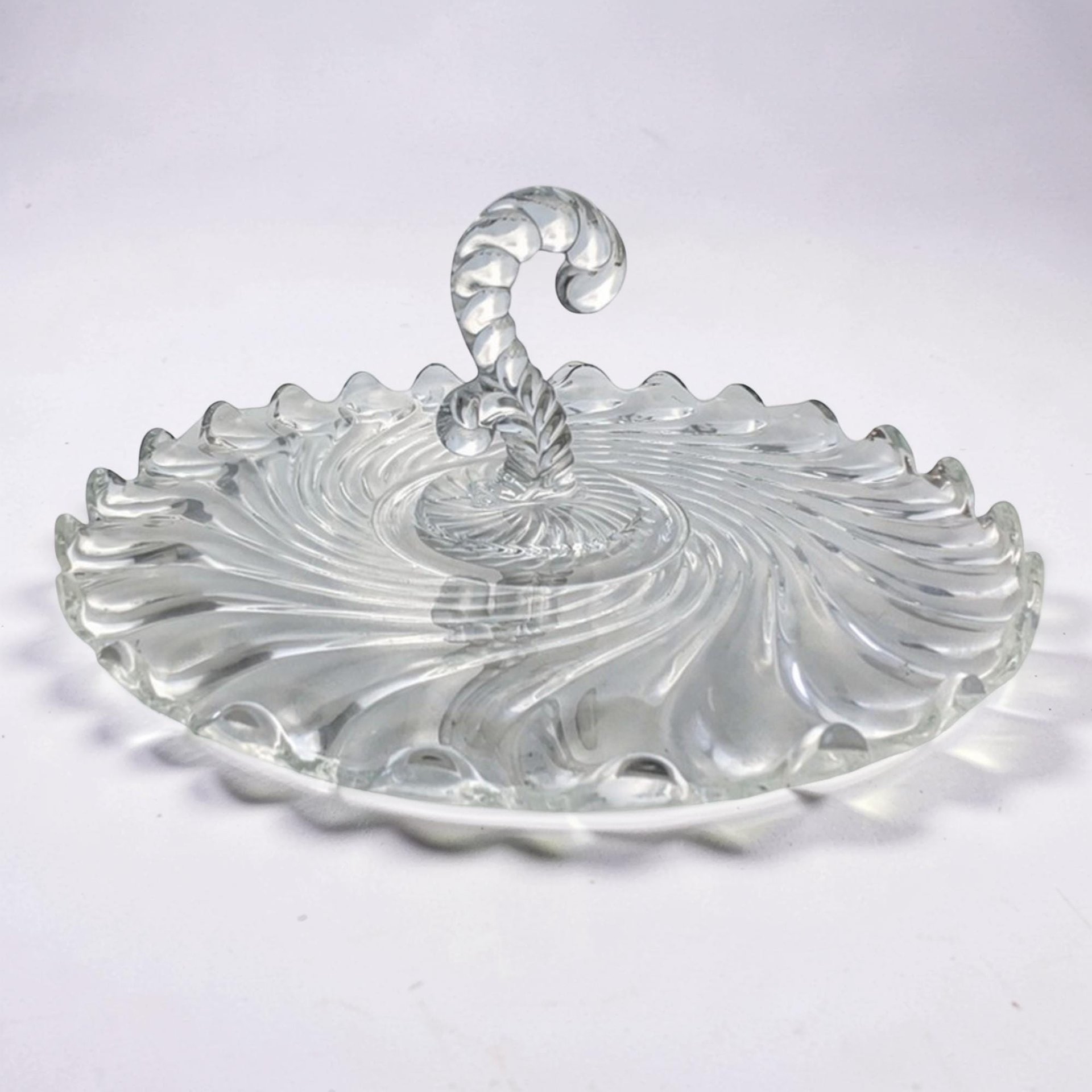 A clear circular glass serving tray with a prominent center handle. It features a dramatic swirling ribbed texture radiating from the center to the scalloped edge. The glass is thick, transparent, and highly reflective.