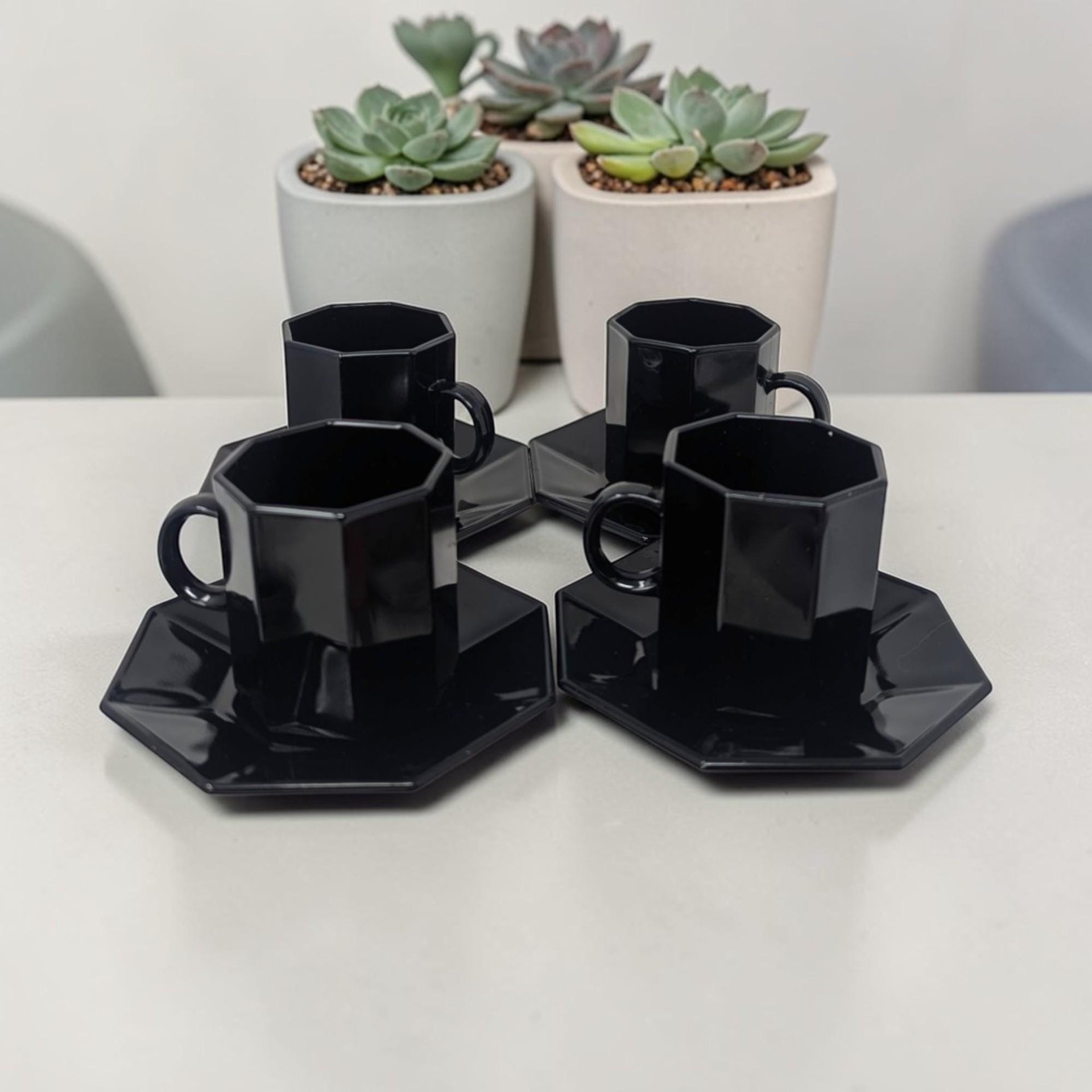 Four small black glass coffee cups and matching saucers. The pieces feature a distinct octagonal geometric shape with a high-shine, opaque black finish. The cups have a small side handle and flat angular sides.