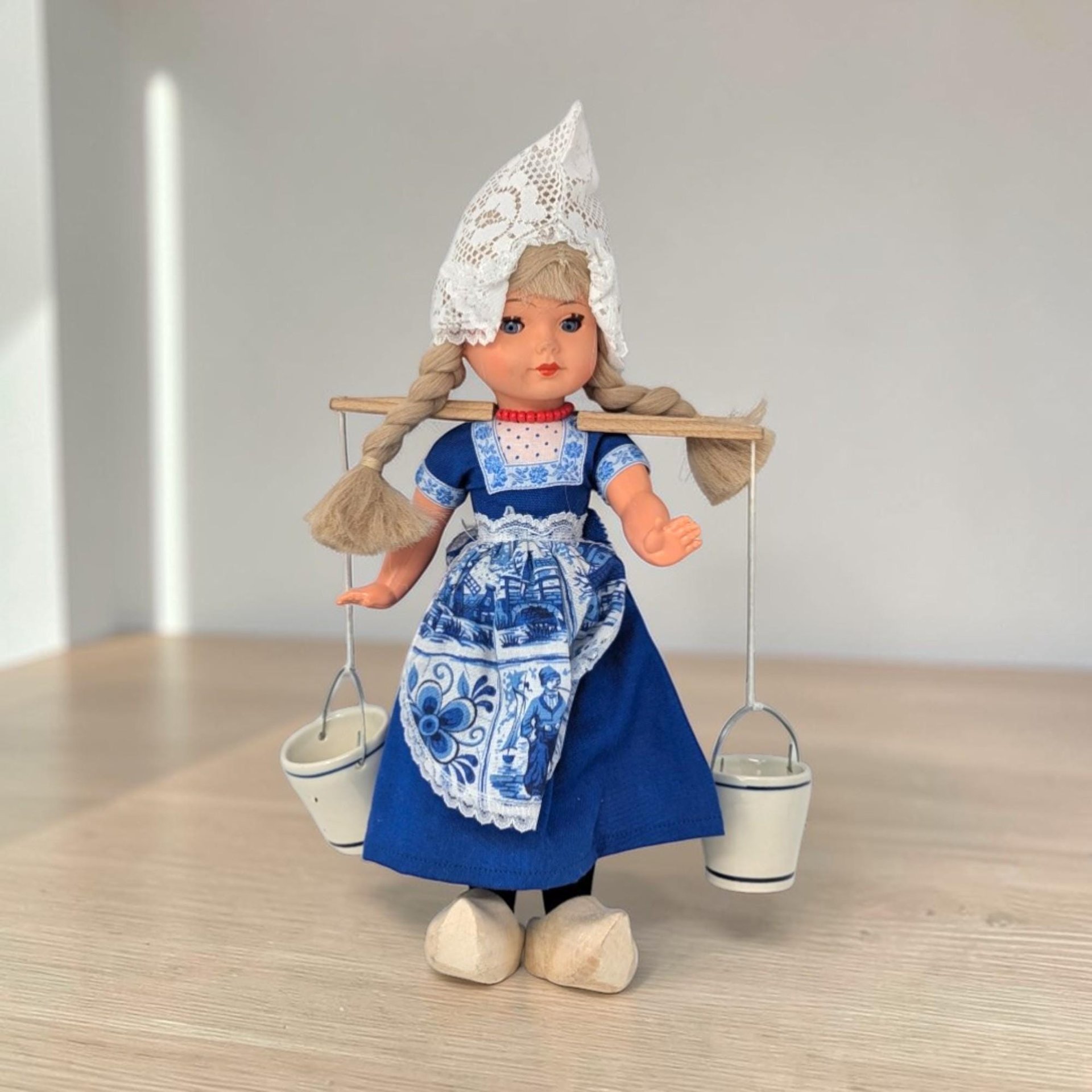A plastic doll wearing a traditional Dutch costume including a pointed white lace cap and blue striped skirt. She carries a wooden yoke across her shoulders with two small blue and white ceramic pails hanging from metal chains.