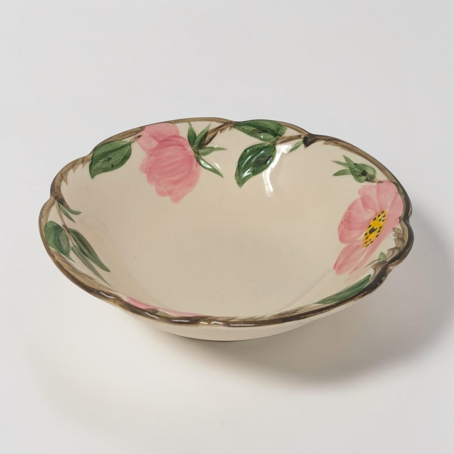 A small circular ceramic bowl with a hand-painted pink desert rose in the center. The scalloped rim is embossed with a brown woody vine and green leaf border on a creamy ivory background. It has a low, shallow profile.