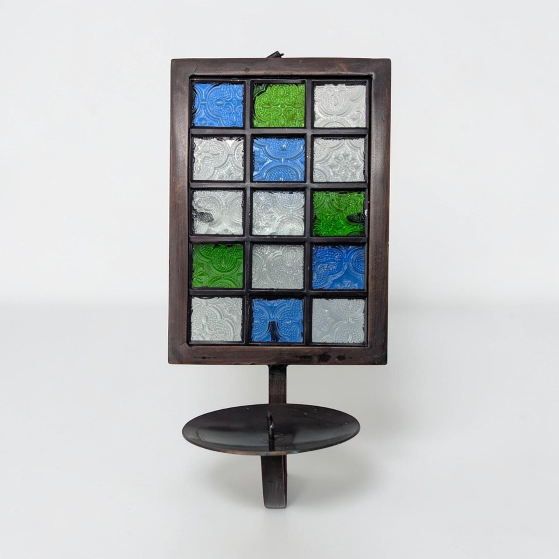 A tall black metal wall sconce featuring a grid of twelve square glass inserts in shades of forest green, cerulean blue, and opaque white. The rectangular frame has a single candle cup at the base and a circular hanging loop.