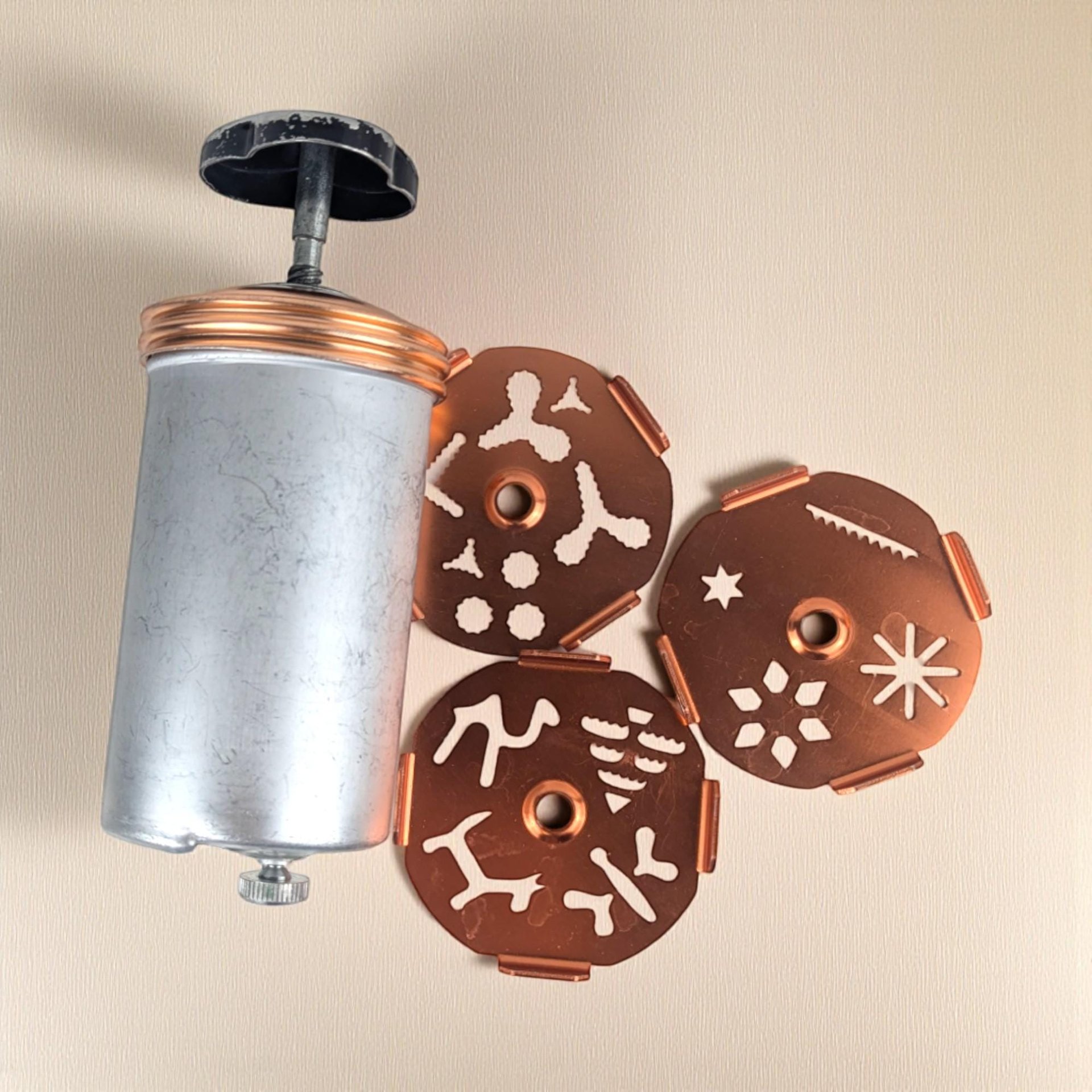 A cylindrical aluminum cookie press with a black knob handle and copper-toned screw cap. Three circular copper-finished template discs with various cutout shapes for dough extrusion lie next to the press. The metal shows a matte, brushed texture.