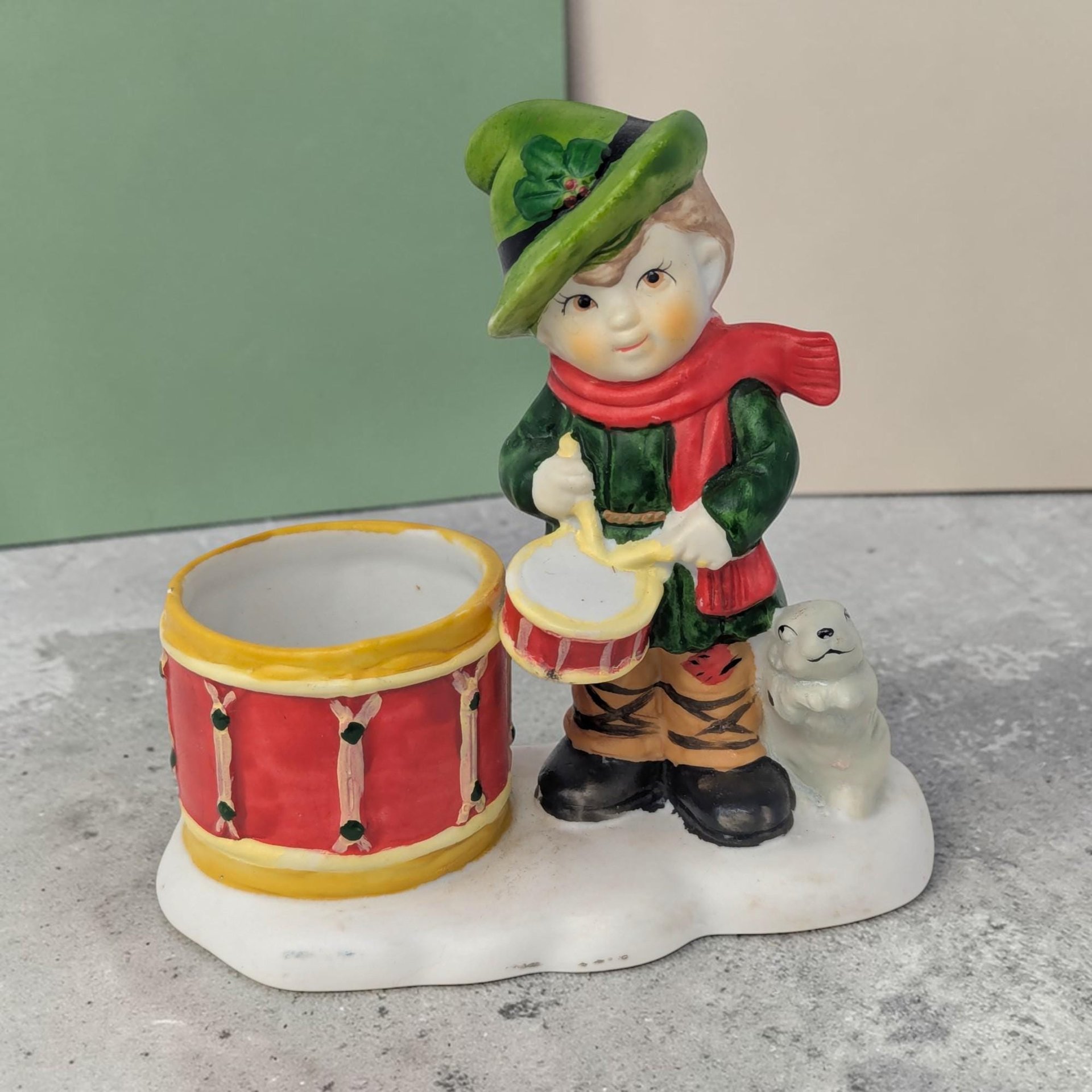 A small ceramic candle holder shaped like a drummer boy in a red and green uniform. The texture is a smooth, matte glaze with a candle holder